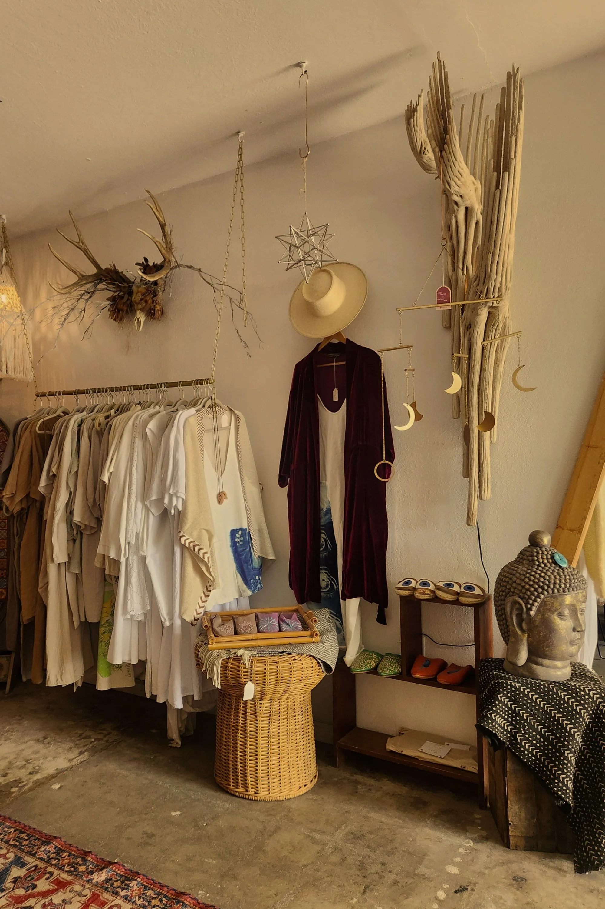 Vintage clothing store interior with hanging garments, decorative items, and rustic decor, featuring a hat, a statue, and woven basket.