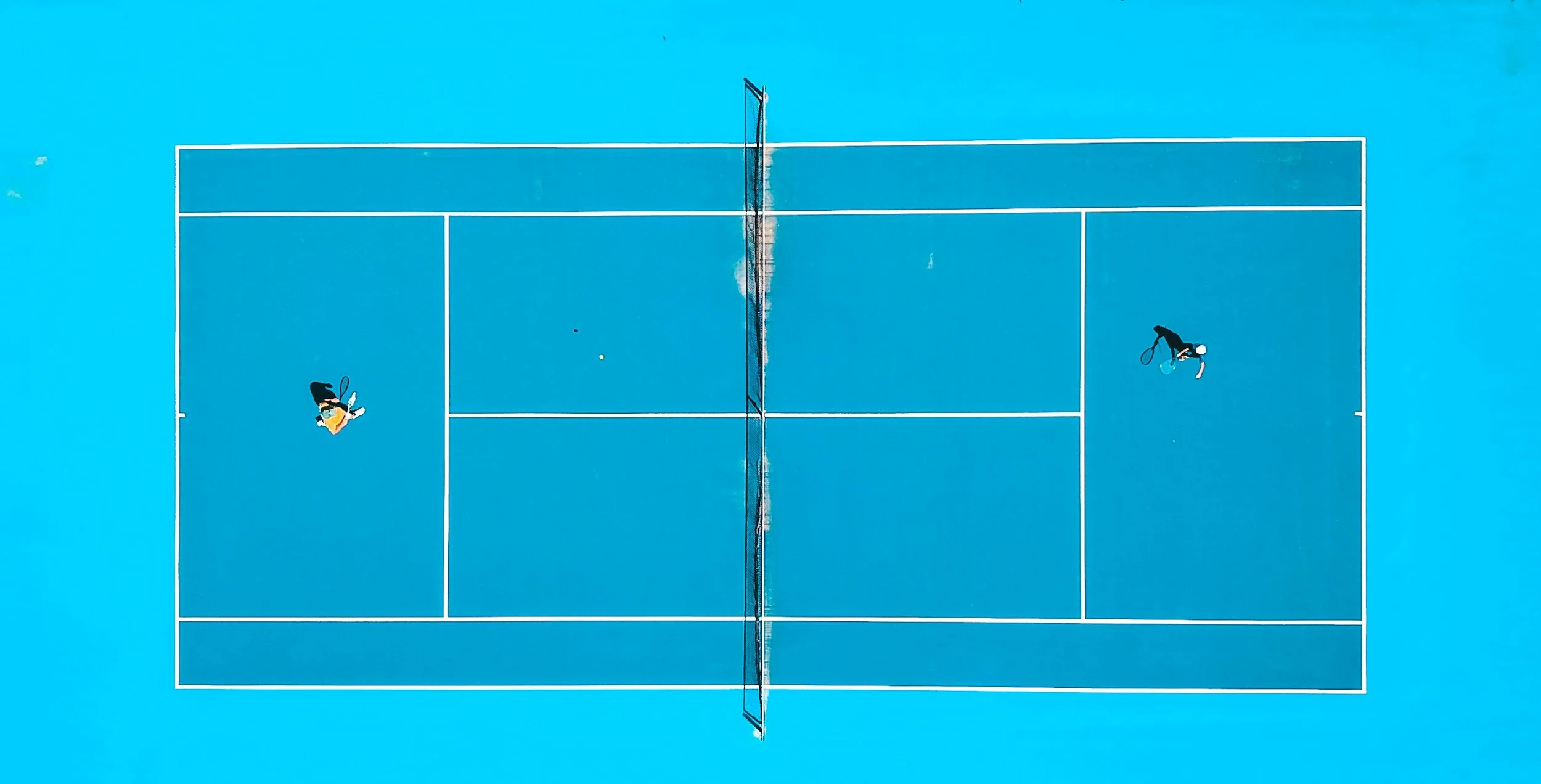 Top-down view of a bright blue tennis court with two players, one on each side of the net, preparing to serve or return a shot.