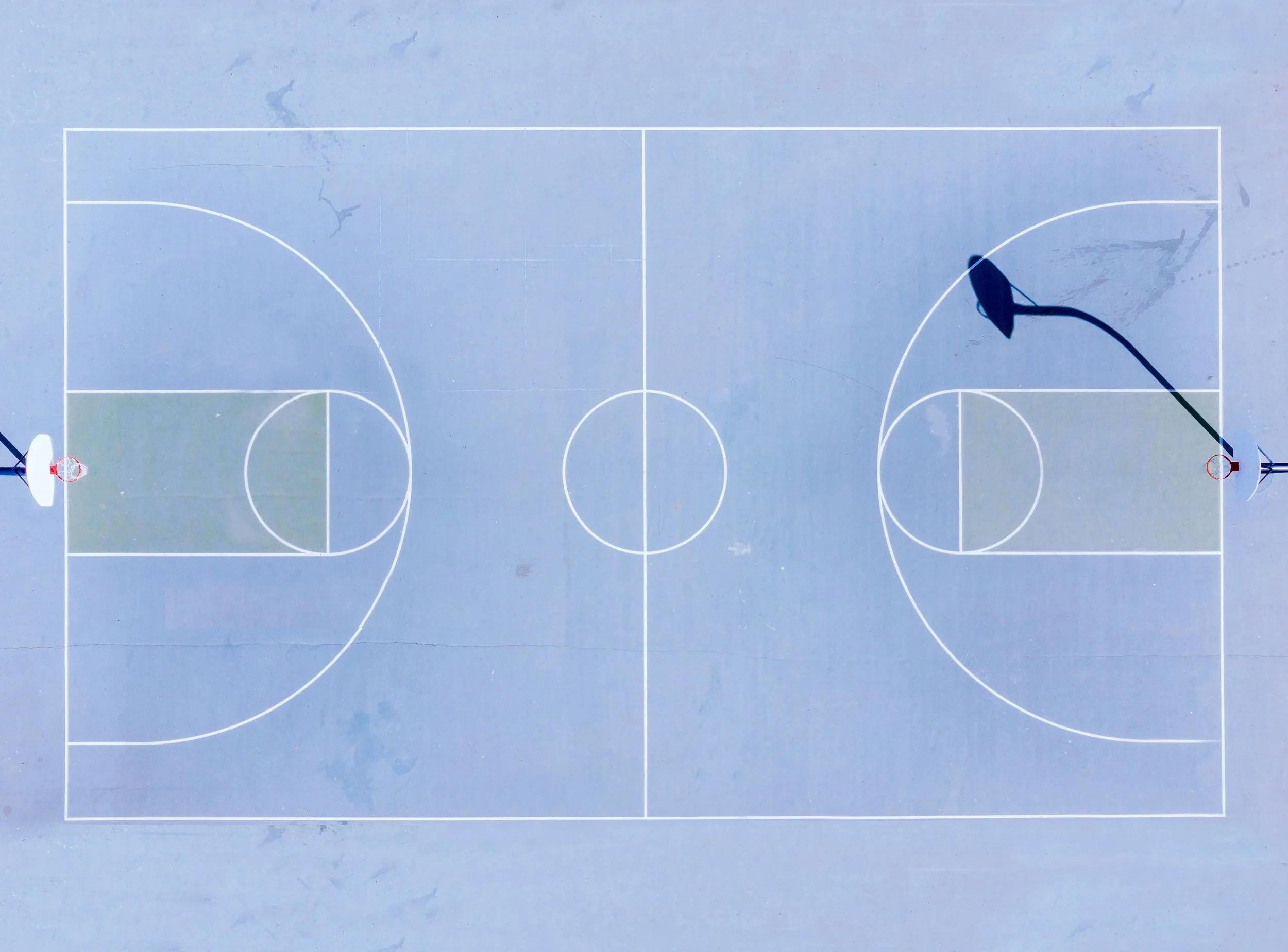An outdoor basketball court viewed from above, with two hoops at opposite ends, marked with white lines for the game.