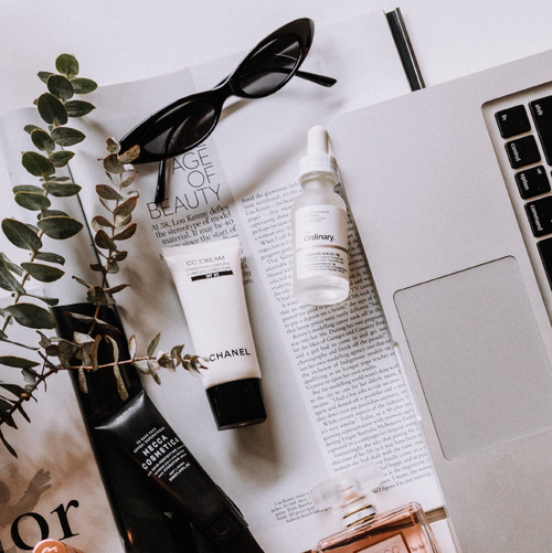 Beauty and skincare products, a pair of sunglasses, a pen, and a laptop on a desk with a plant and magazine pages.