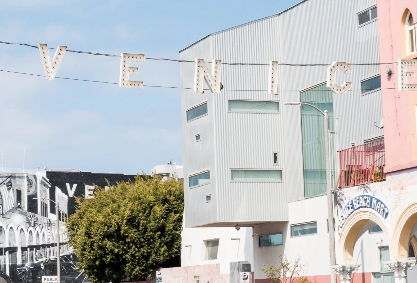 Sign spelling 'VENDING' with individual bulbs in front of a modern building and a colorful building with an arch, Christmas lights, tree, and parking meters.