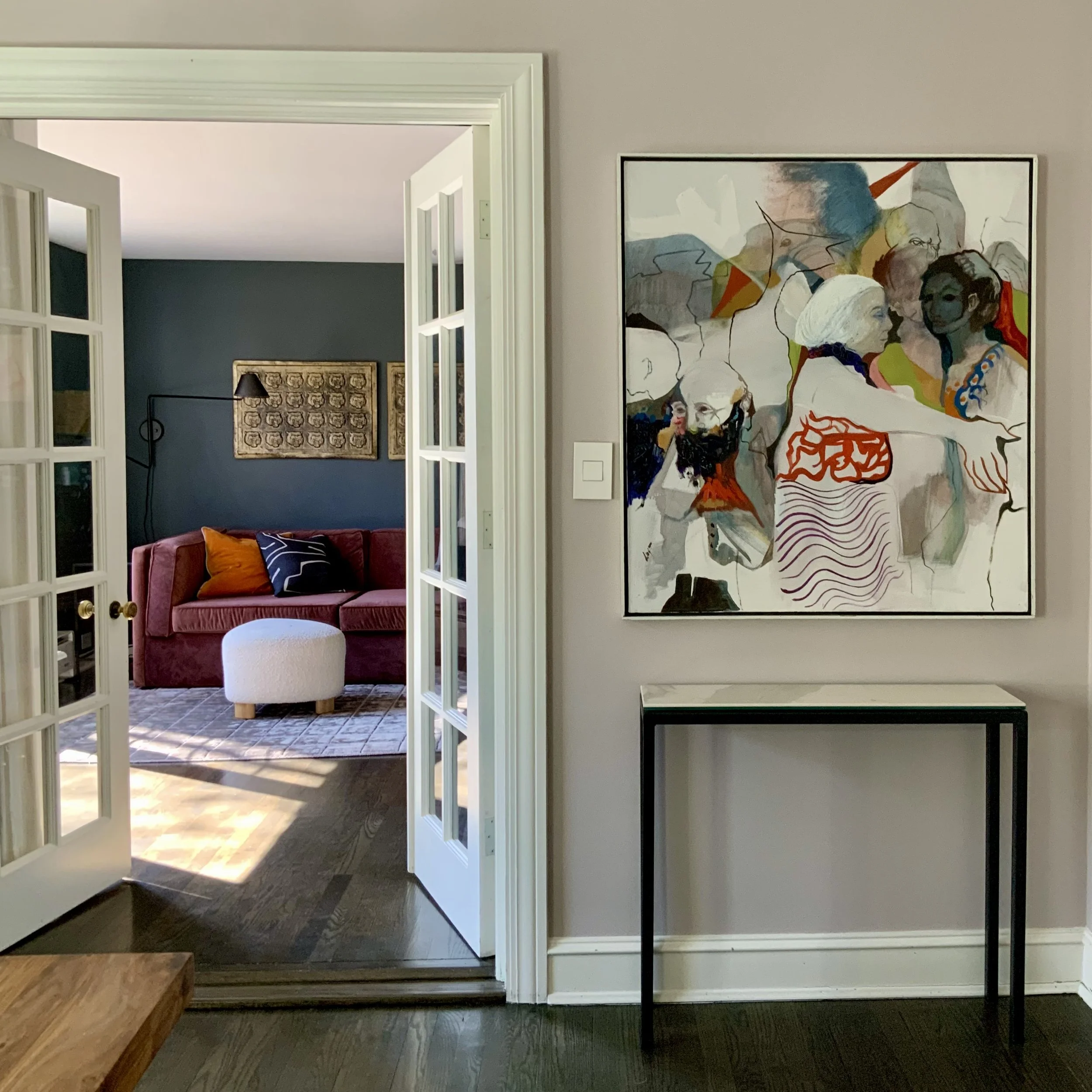 Interior view of a room with French doors leading to a second room featuring a maroon sofa and decorative pillows. A colorful abstract painting hangs on the taupe wall next to a minimalist console table. The floor is dark wood.