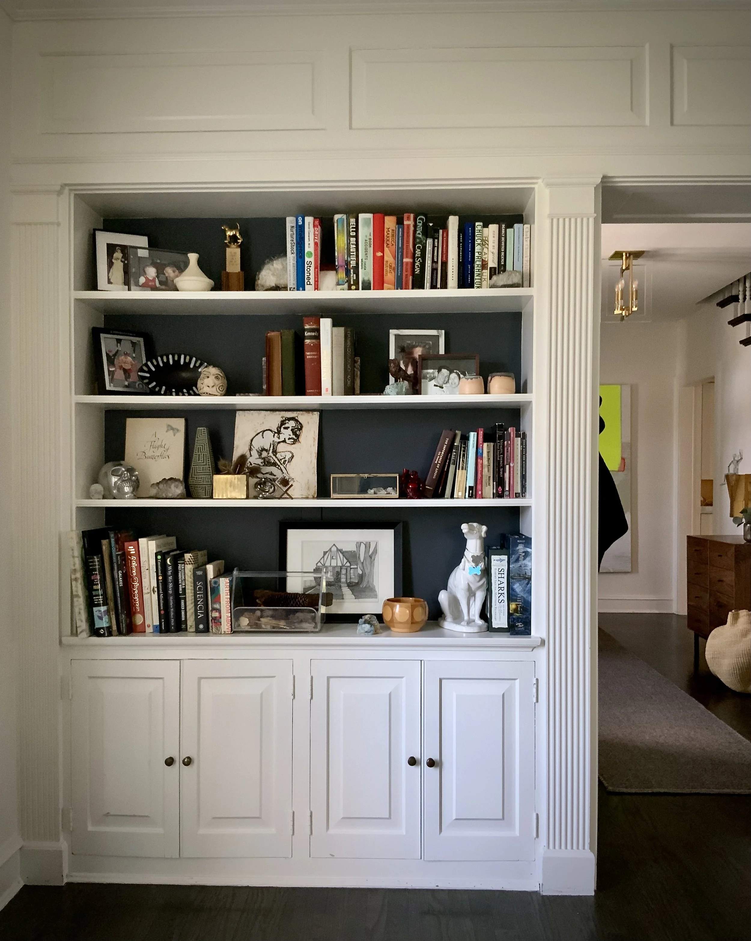 White built-in bookshelf with various books, framed photos, small statues, and decorative items. Lower section has closed cabinets.