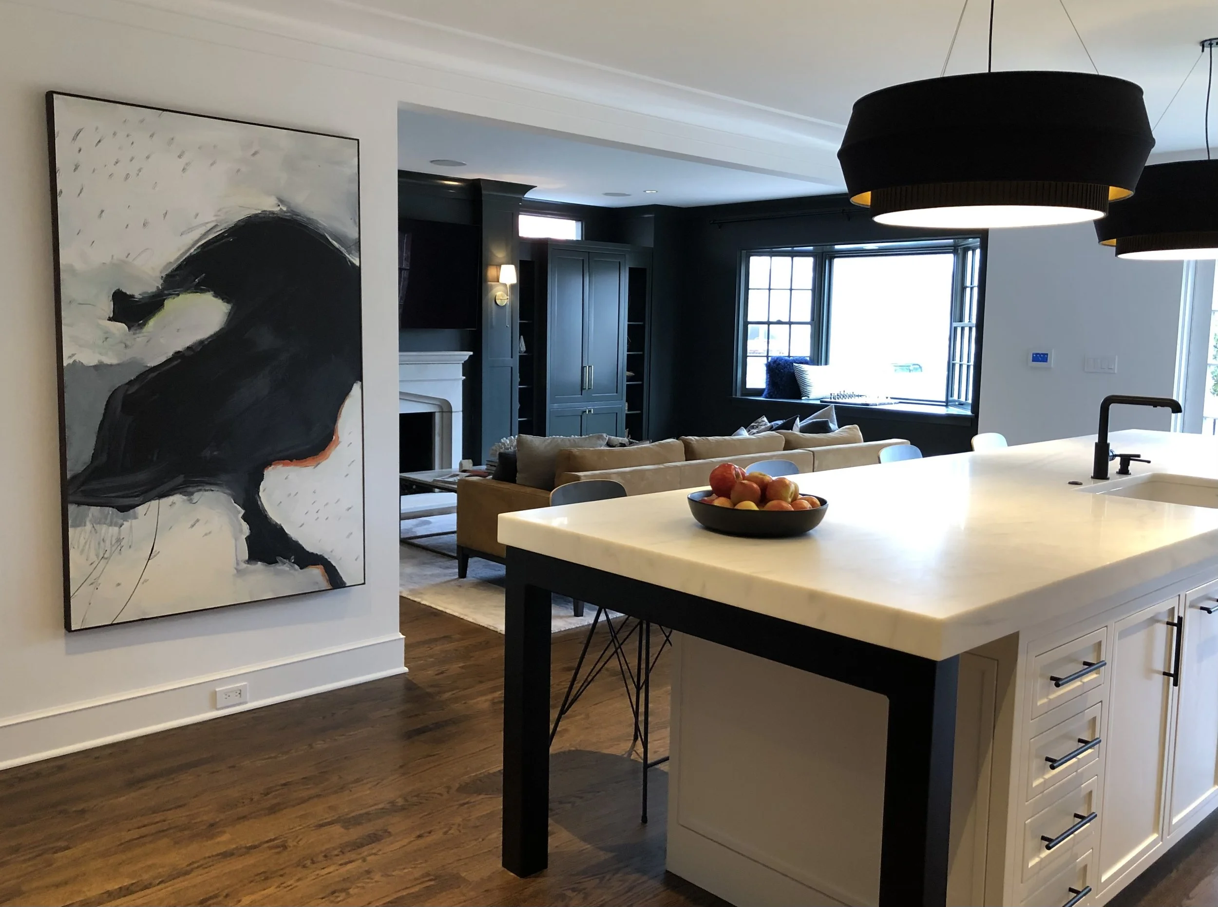 Contemporary open-concept kitchen and living area with modern art on wall, marble island, black pendant lights, and a bowl of fruit.