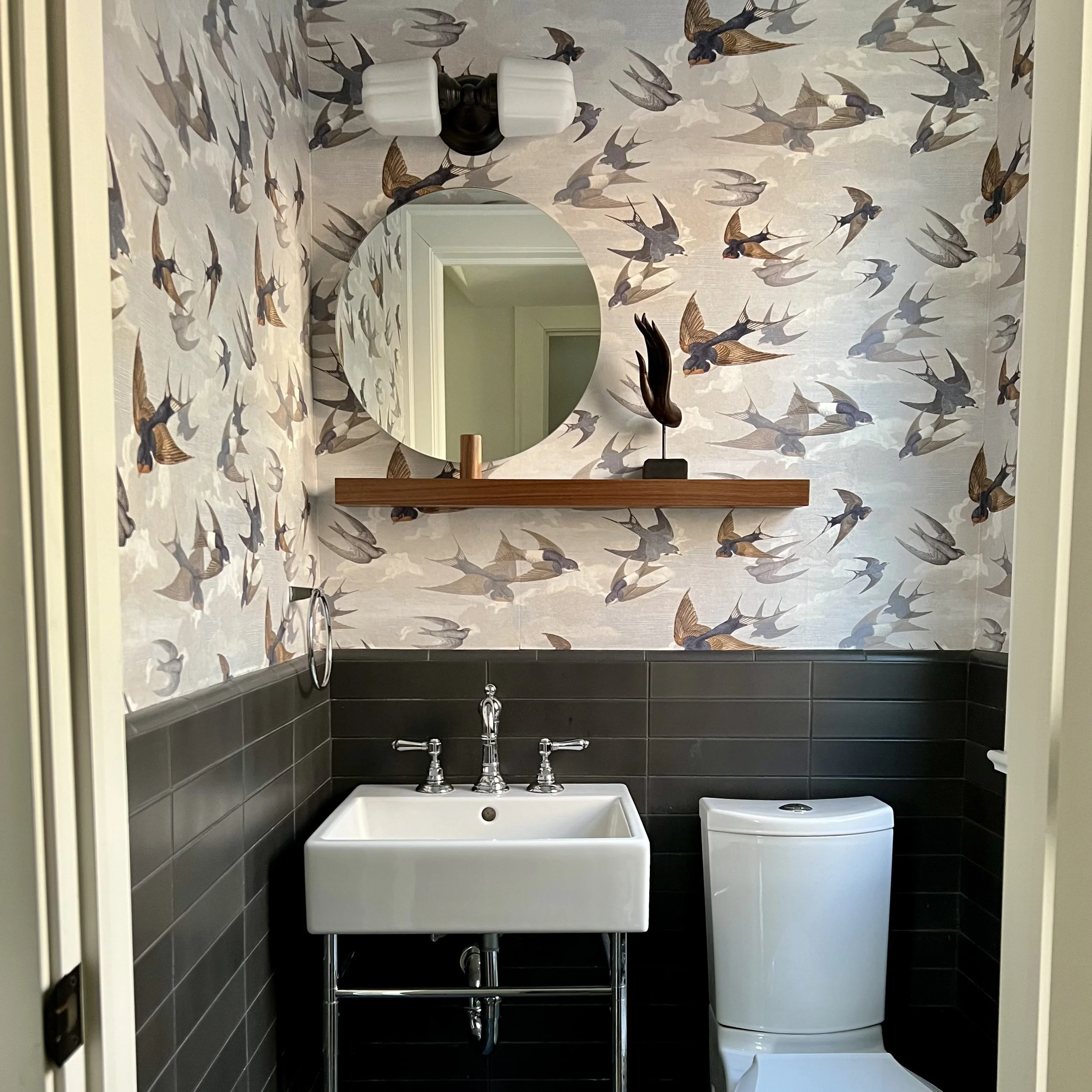 Small bathroom with bird-patterned wallpaper, round mirror, white sink, toilet, and dark tiles.