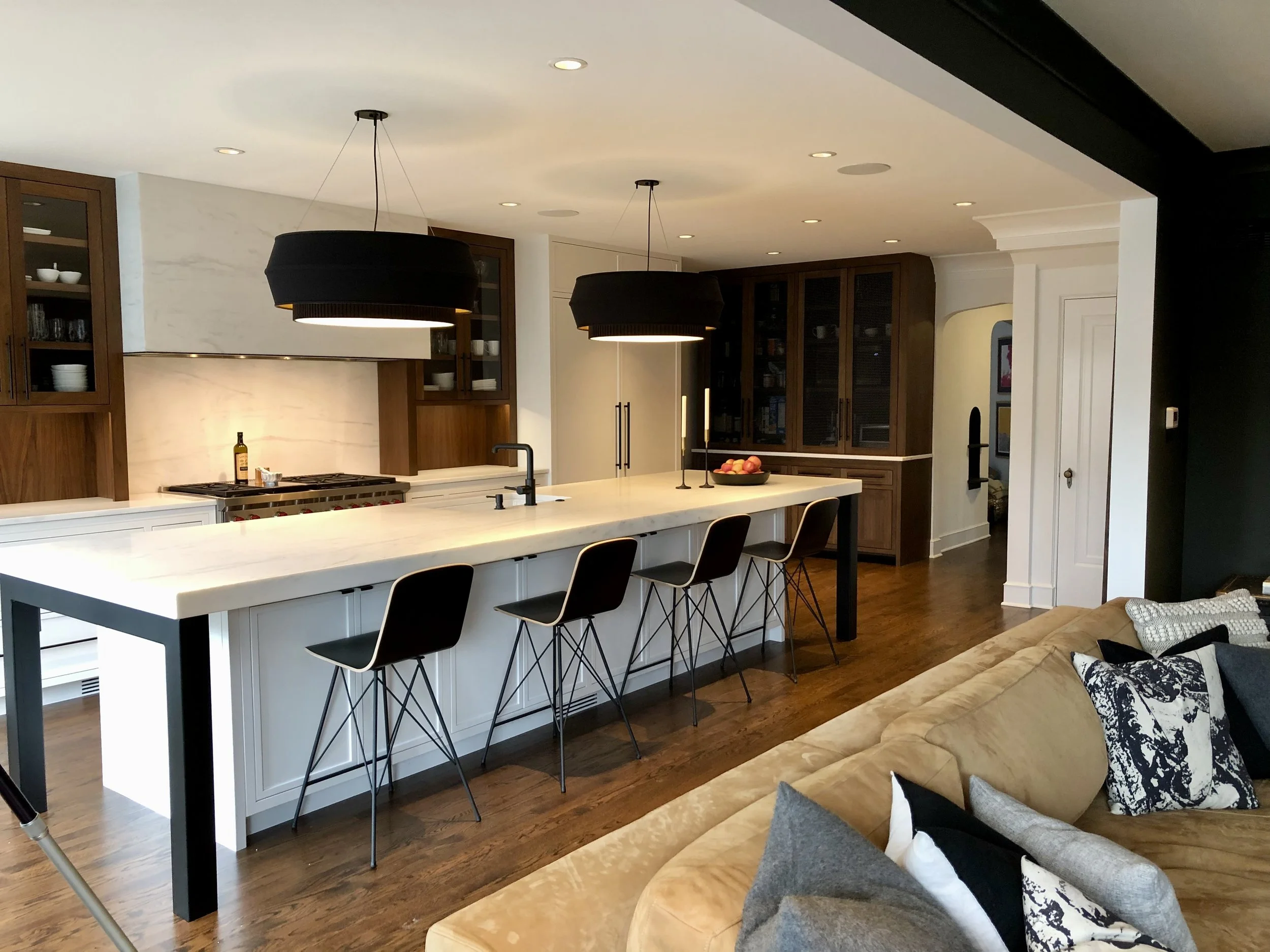 Modern kitchen and living area with a large white island, black chairs, overhead pendant lights, dark wood cabinets, and beige sofa.