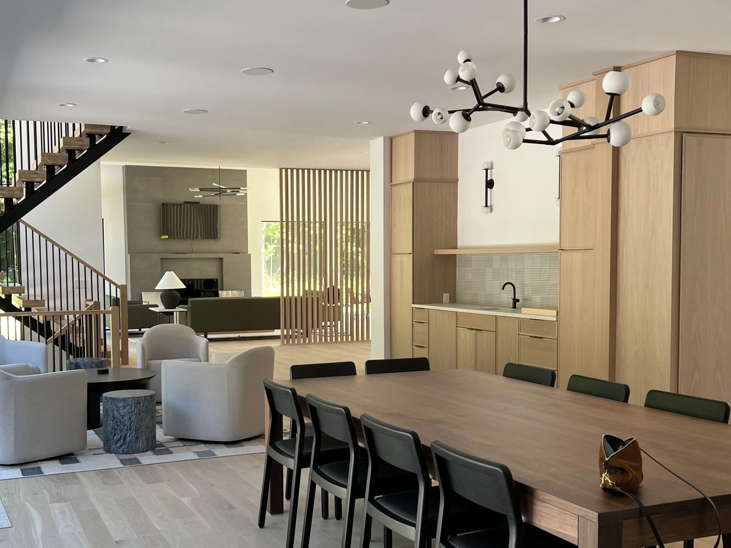 Modern open-concept living and dining room with minimalist design, featuring a large wooden dining table, contemporary chandelier, beige armchairs, wooden partition, and a staircase with black railing.