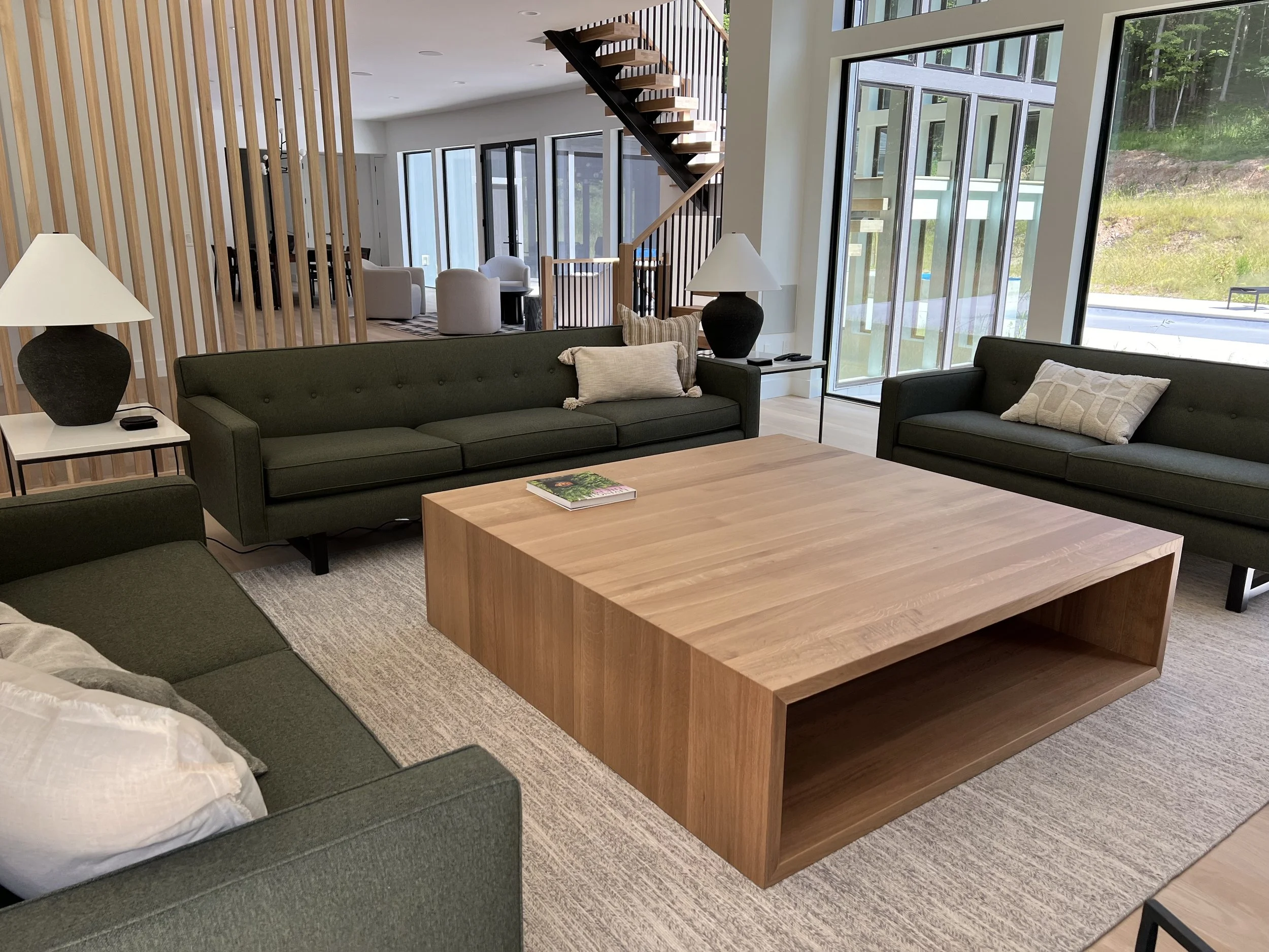 Modern living room with green sofas, a large wooden coffee table, table lamps, and large windows with natural light.