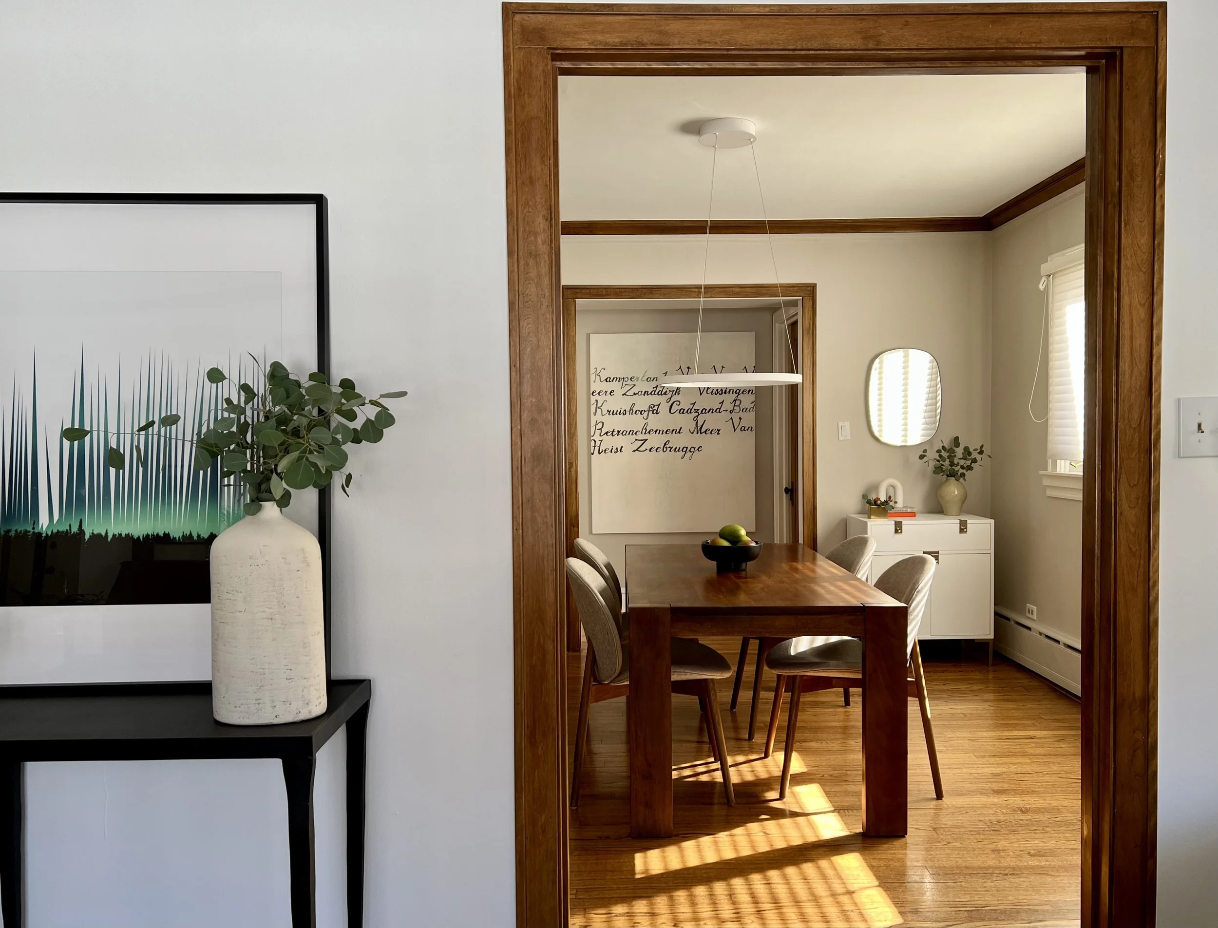 Interior view of a dining room with a wooden table, four chairs, a wall mirror, artwork, and decorative plants on side tables.