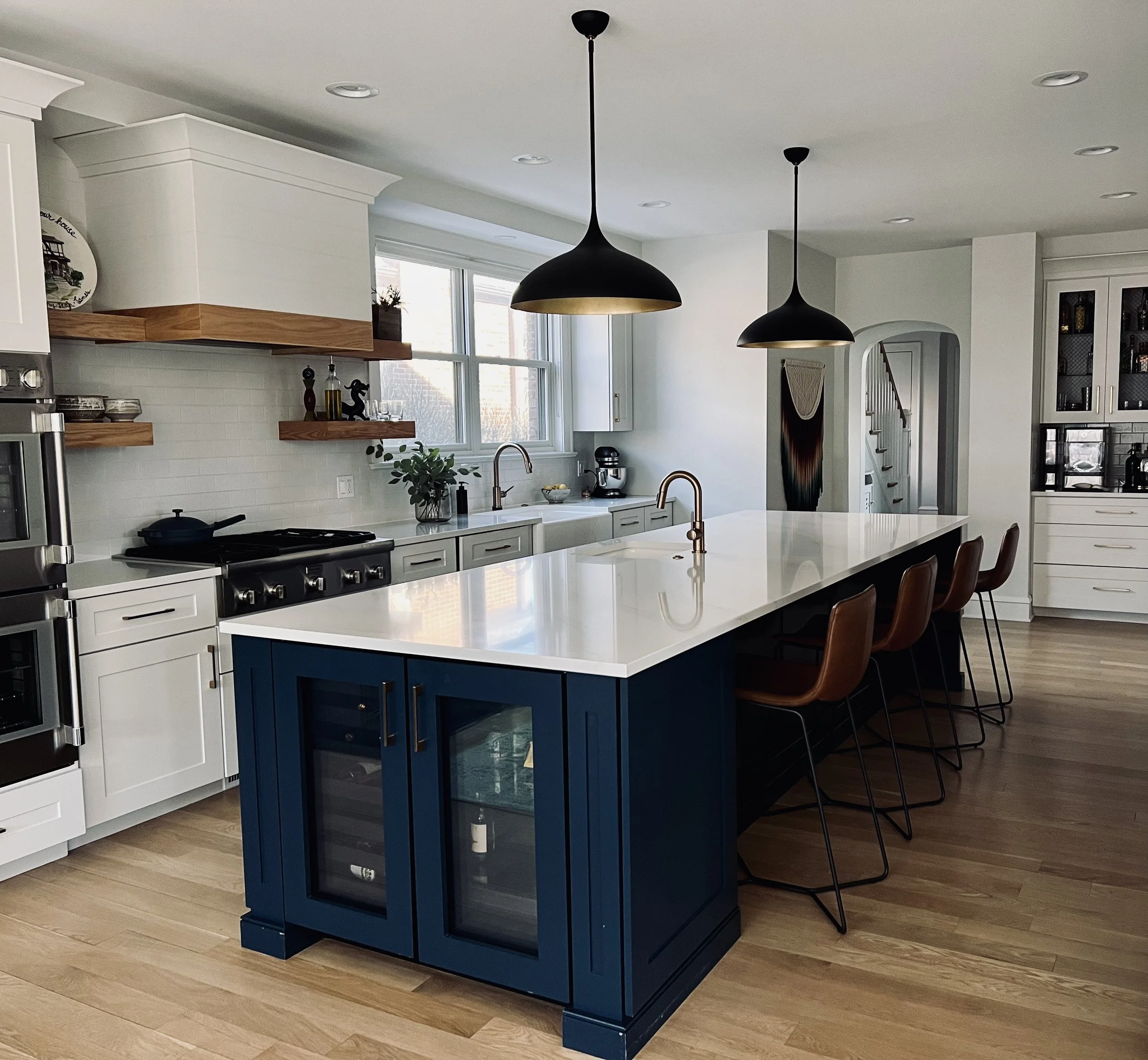 Modern kitchen with large white island, blue base cabinets, brown bar stools, pendant lights, stainless steel appliances, and wood flooring.