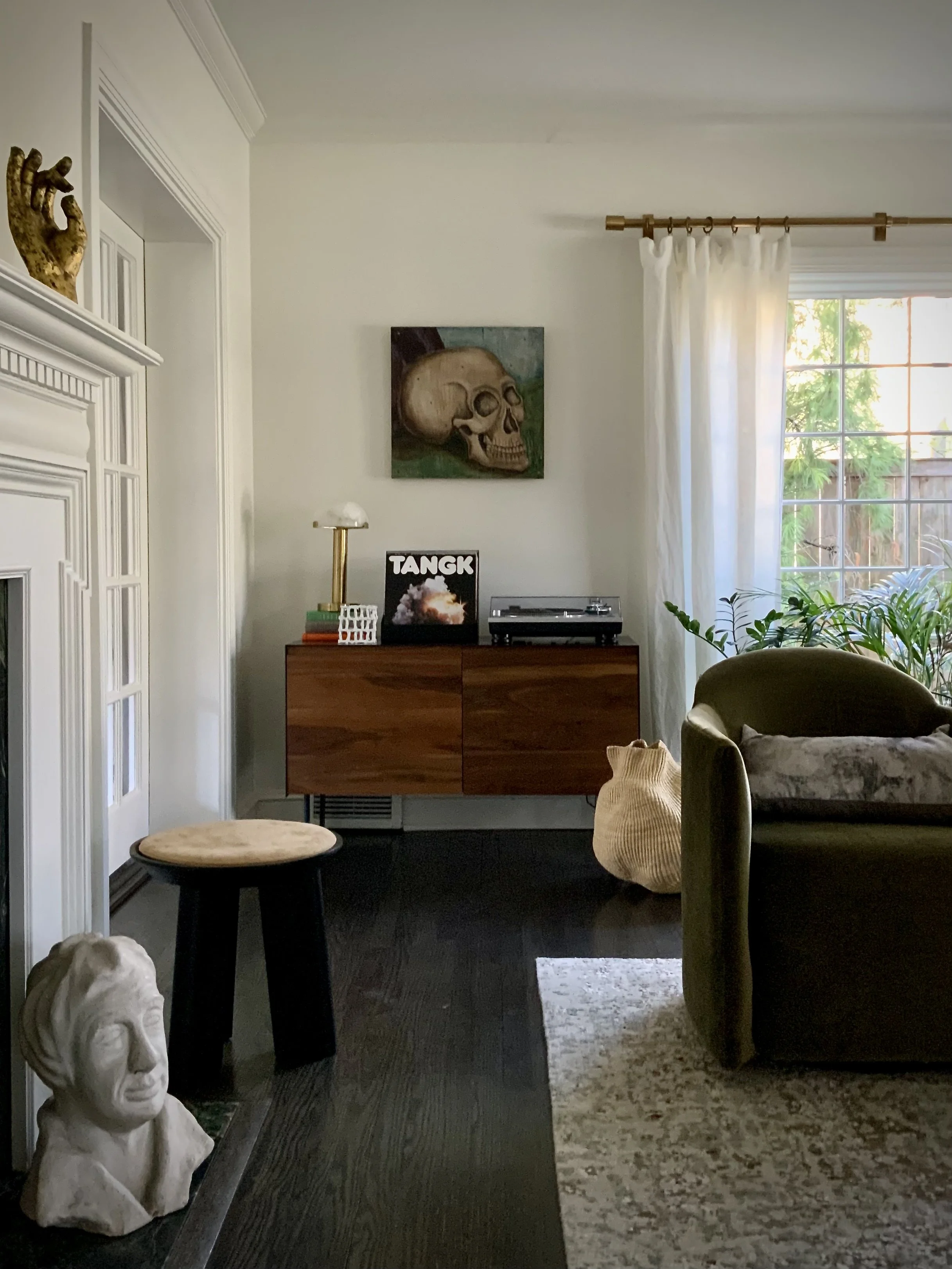 Modern living room with a white wall featuring a skull painting. A brown wooden sideboard holds a lamp, a vinyl record, and a record player. There's a plush green armchair, a white curtain on a window, and a decorative basket on the floor. A statue's