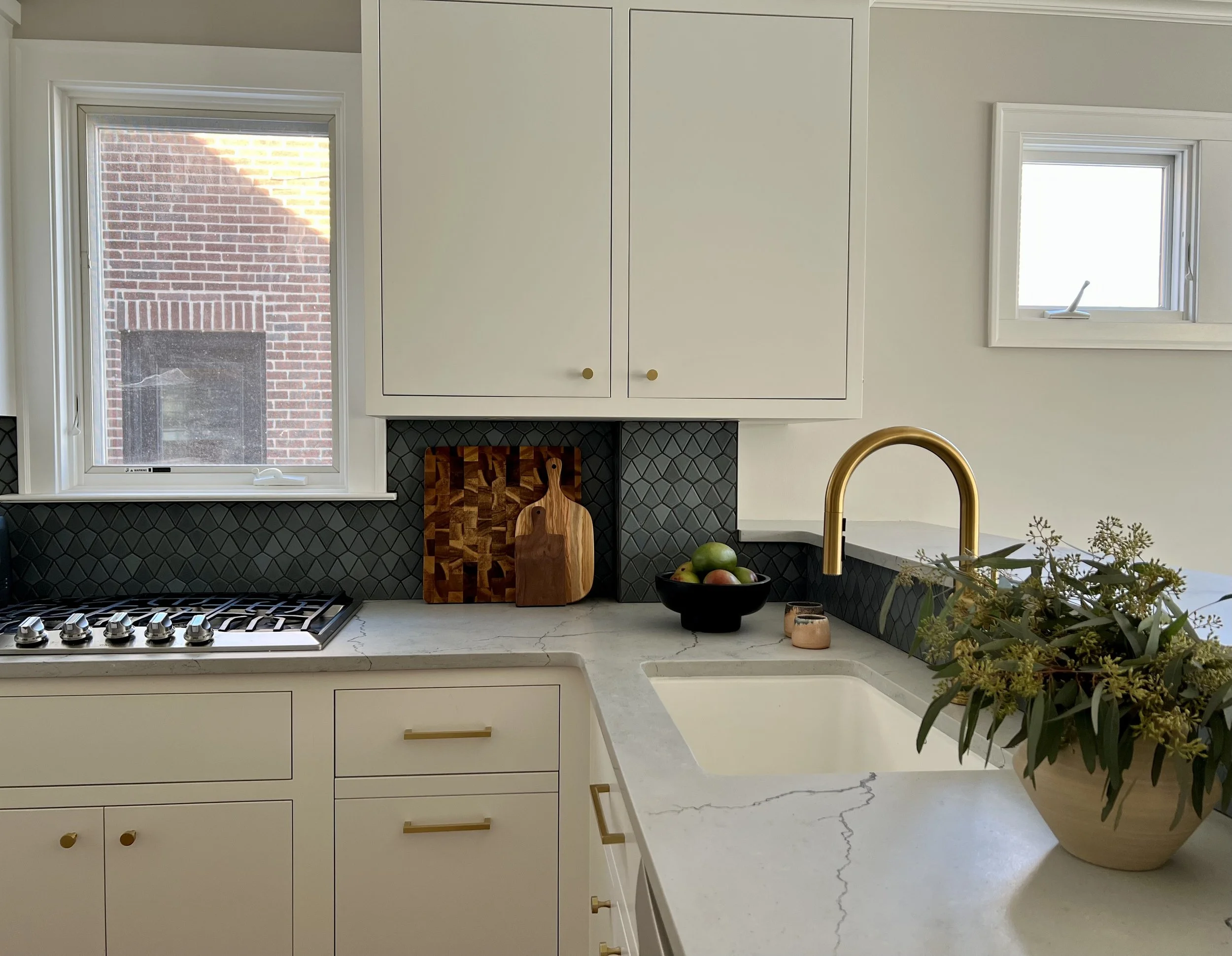Modern kitchen with white cabinets, gold faucet, black backsplash, cutting boards, and a bowl of fruit.