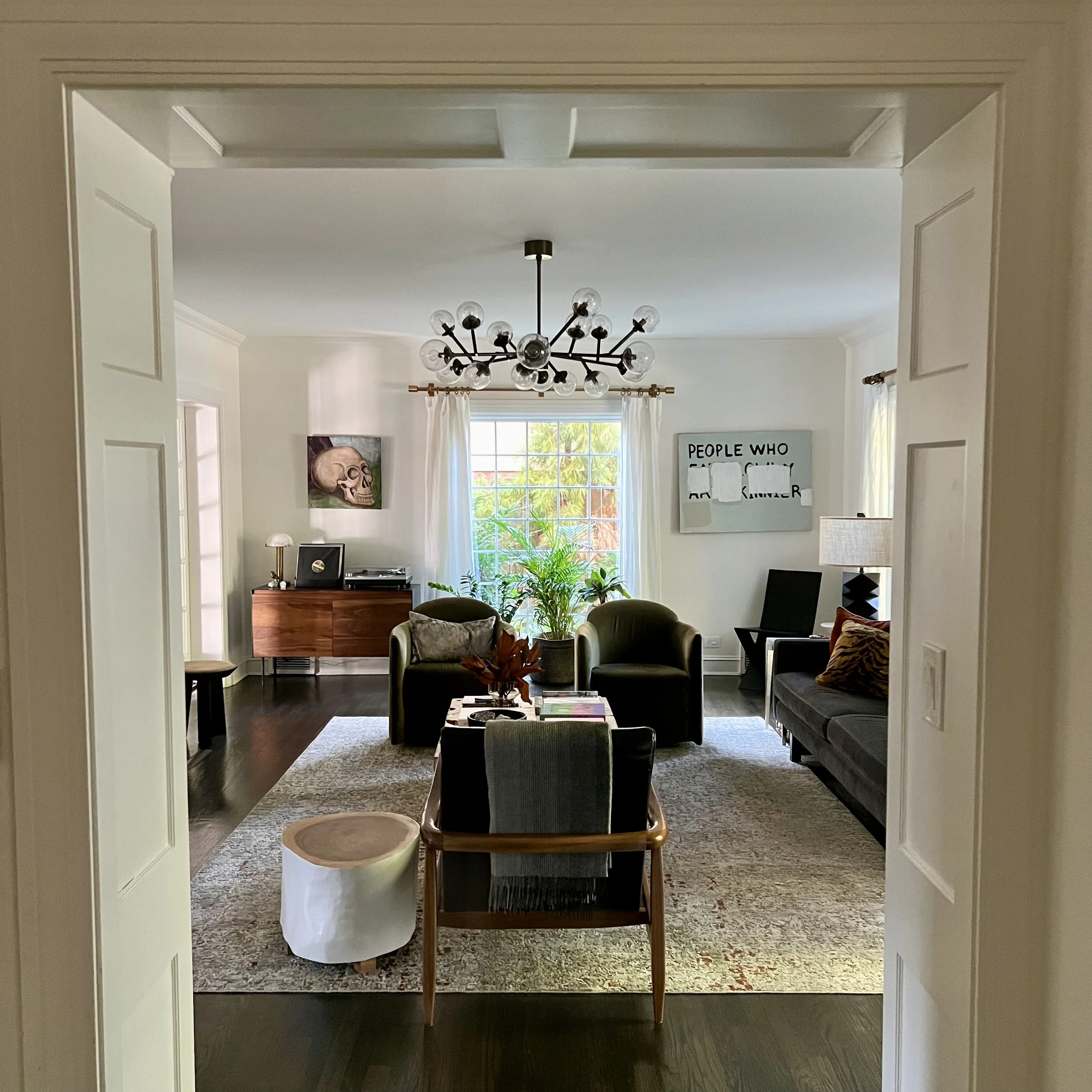 Modern living room with contemporary chandelier, dark wood floors, and neutral furniture. Features a gray sofa, green armchairs, coffee table, side console, wall art, and window with natural light.