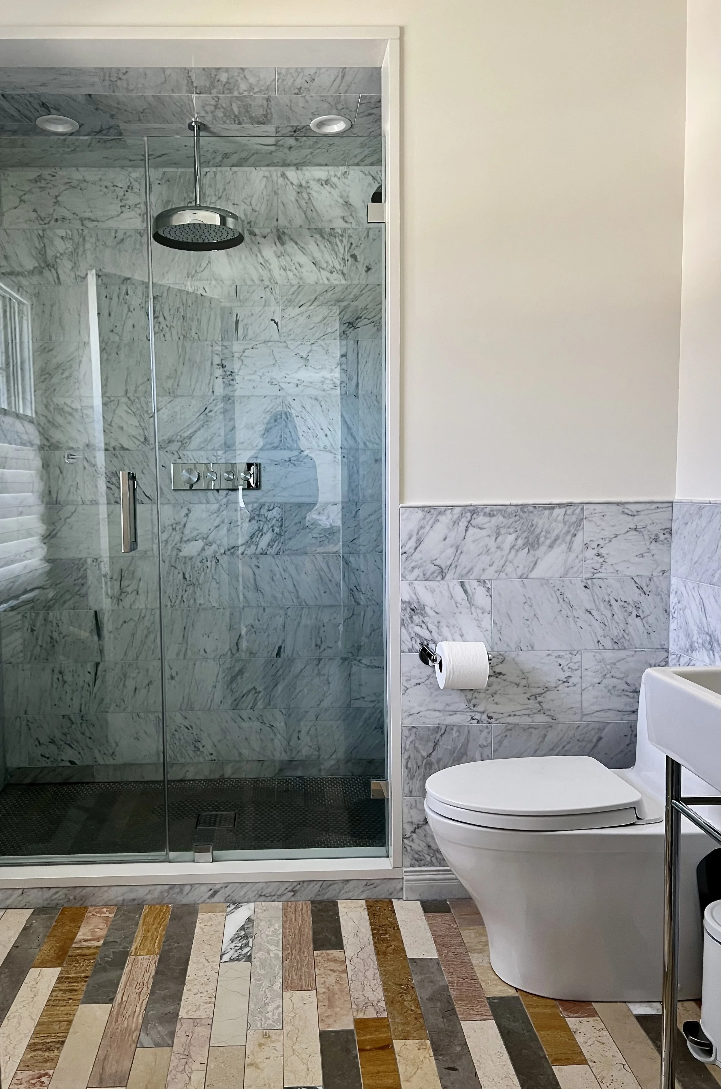 Modern bathroom with marble tile walls, walk-in glass shower, rain showerhead, white toilet, and colorful tile floor.