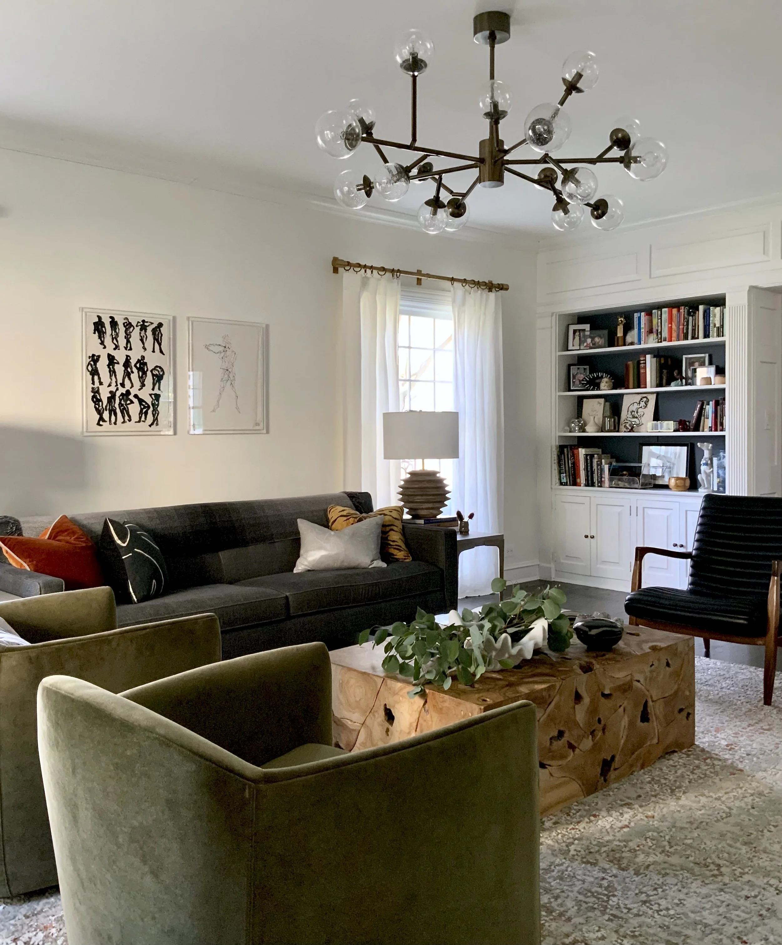 Modern living room with a dark gray sofa, green armchairs, wooden coffee table, and wall art. A chandelier hangs above, with a bookcase in the background.