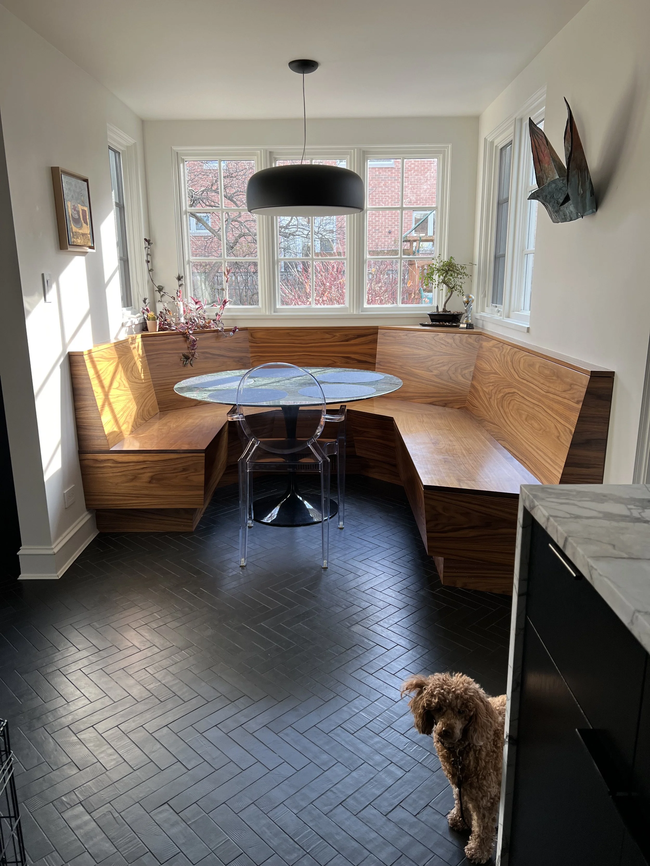 A modern dining nook with a wooden bench seating, a round glass table, and a transparent chair. A black pendant light hangs above. The space has large windows and decorative accents. A small dog is visible on the black herringbone-tiled floor.