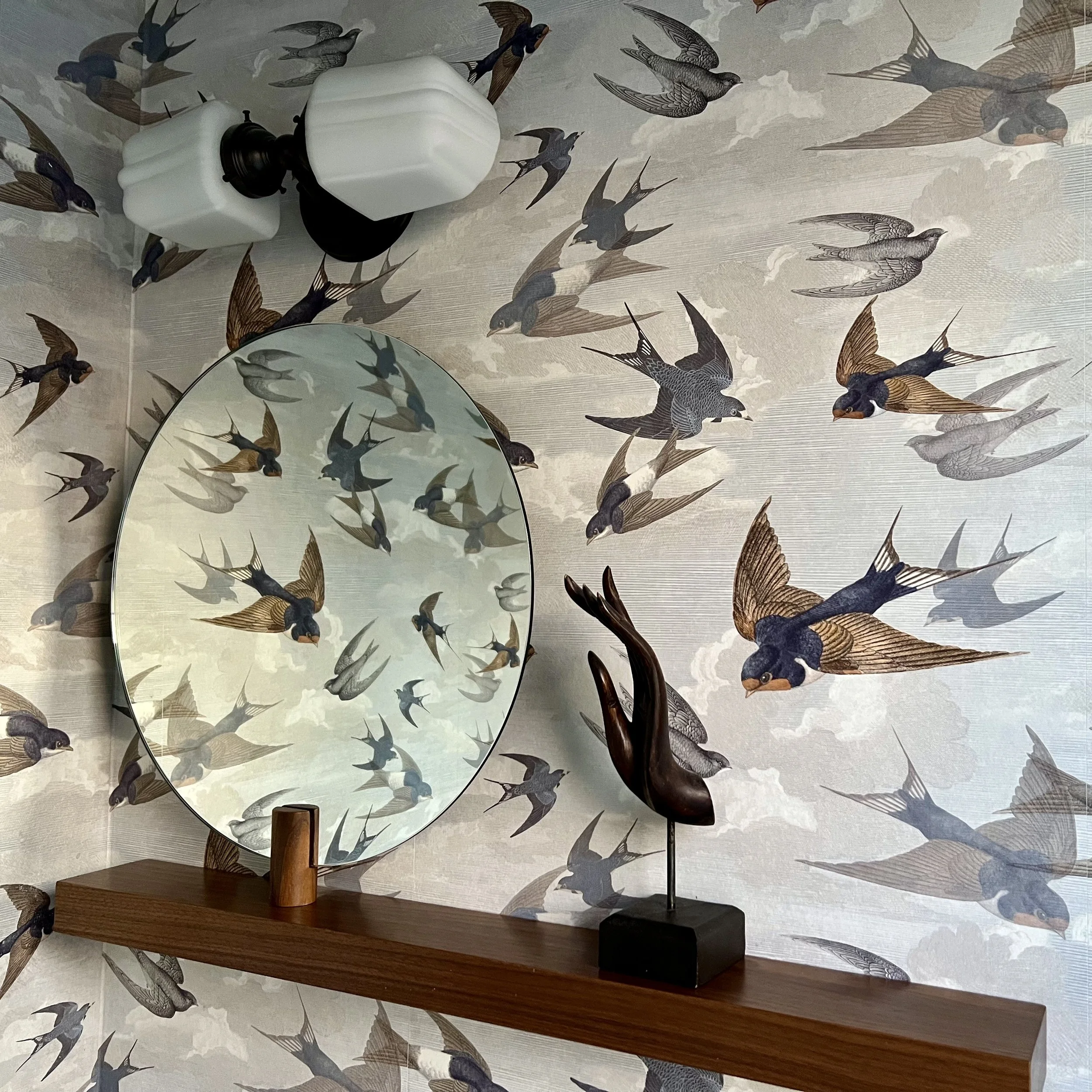 Room with bird-themed wallpaper, round mirror, wall sconce, wooden shelf, and carved bird sculpture.