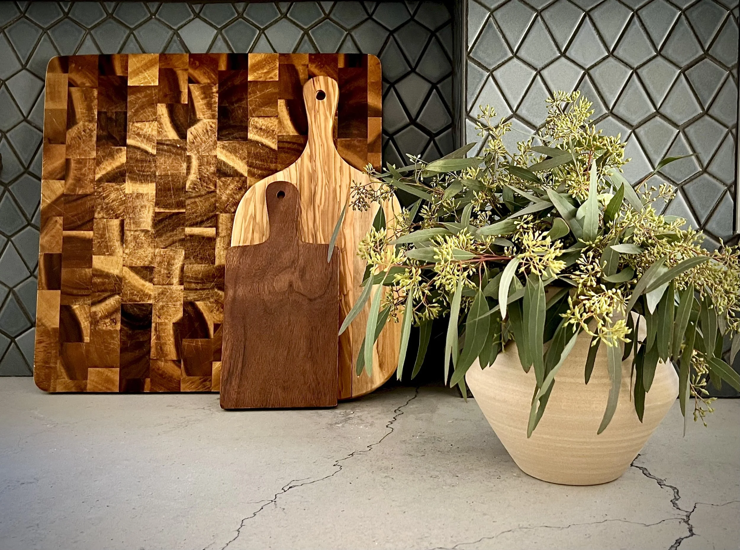 Three wooden cutting boards of varying sizes and wood types are displayed upright against a tiled backsplash. Next to them is a decorative ceramic pot filled with green plants and yellow flowers.