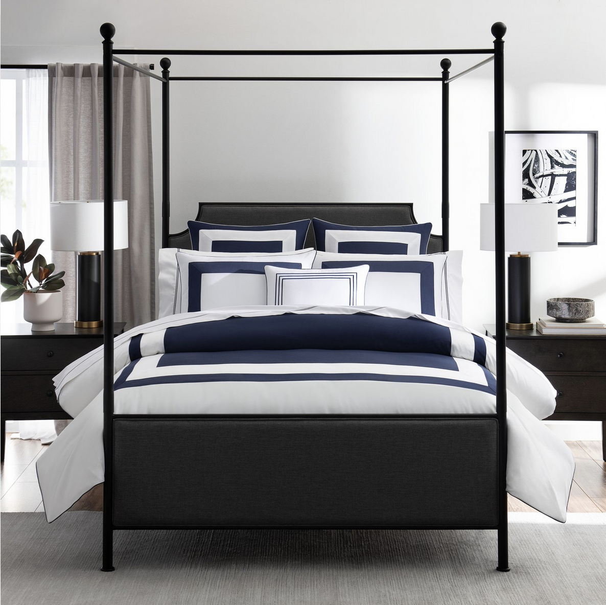 A modern bedroom with a black four-poster bed, navy and white striped bedding, two bedside tables with matching lamps, and abstract art on the wall.