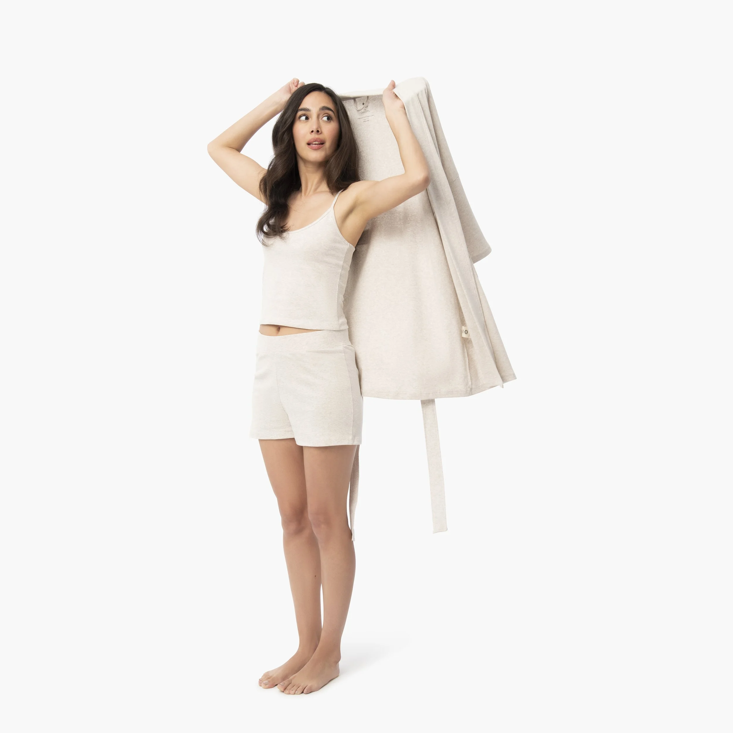 Young woman standing barefoot, holding a beige coat on her shoulder and looking to the side, wearing a matching beige sleeveless top and shorts against a white background.