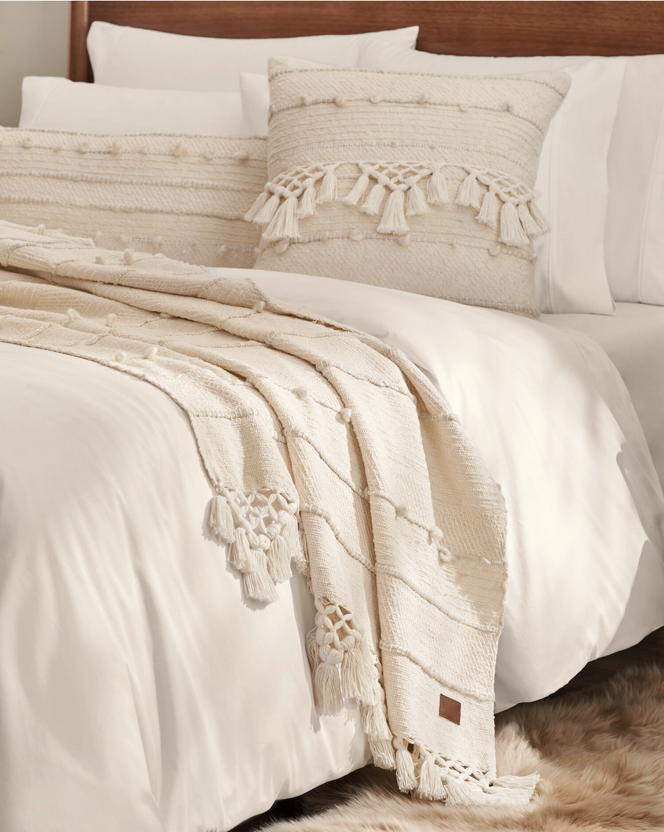 A neatly made bed with white sheets, a beige textured throw blanket with tassels, and matching decorative pillows with tassel accents. A soft, furry rug is partially visible on the floor.