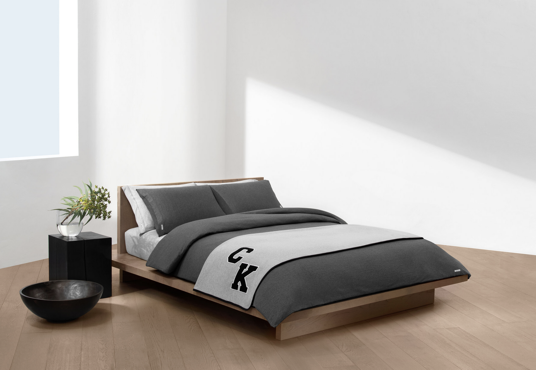 Minimalist bedroom with a low wooden bed frame, gray and black bedding, a Potter-style black bowl on the light wood floor, a black nightstand with a potted plant, and sunlight casting shadows on a white wall.