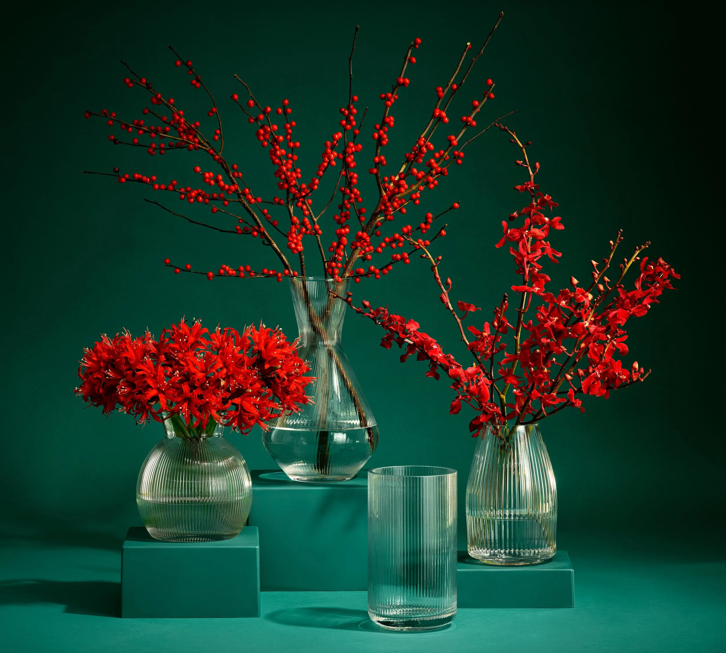 Arrangement of three vases with vibrant red flowers and berries, set against a dark green background.