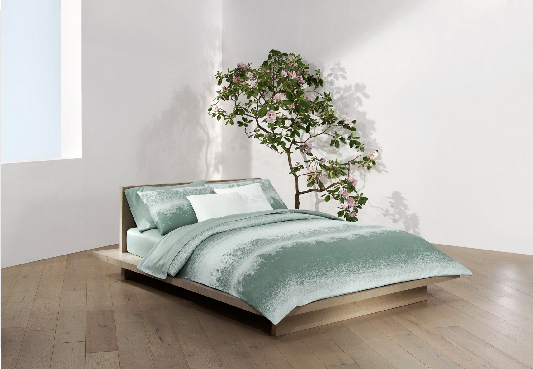 A bedroom with a low wooden bed, green bedding, and a large pink-flowered tree shadow on white walls.