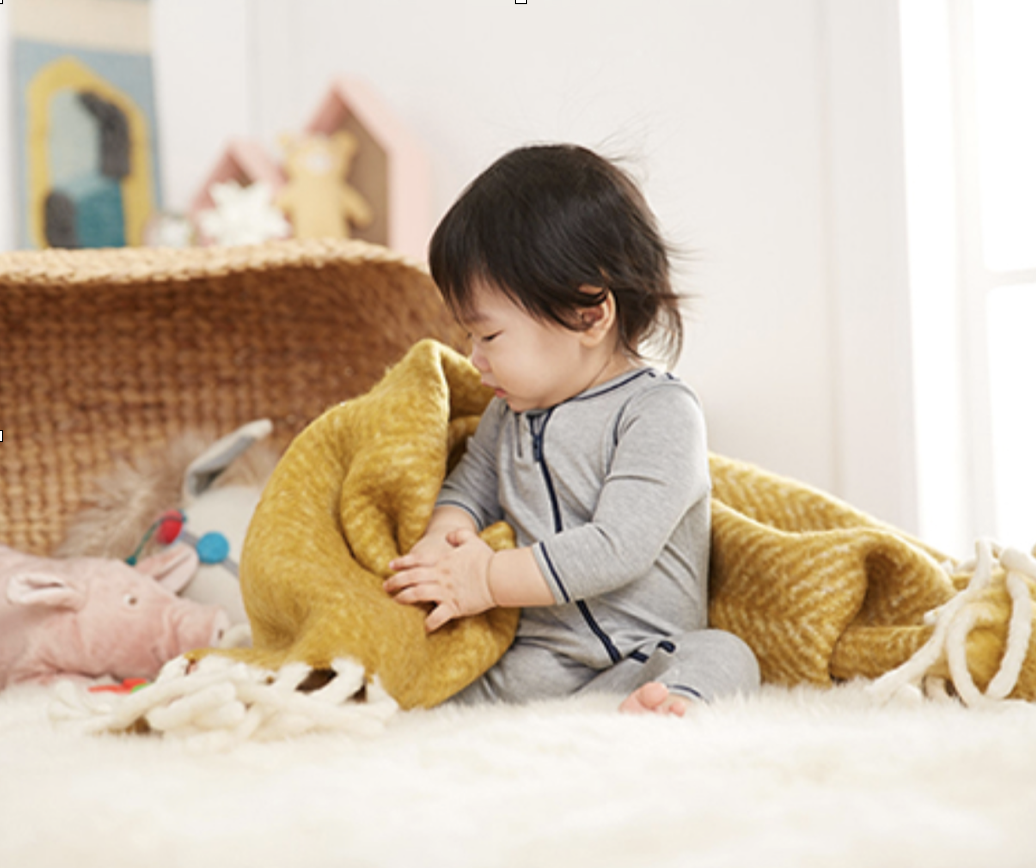 Young child sitting on a fluffy white rug, holding and hugging a yellow stuffed animal in a cozy room with toys and decorative items in the background.