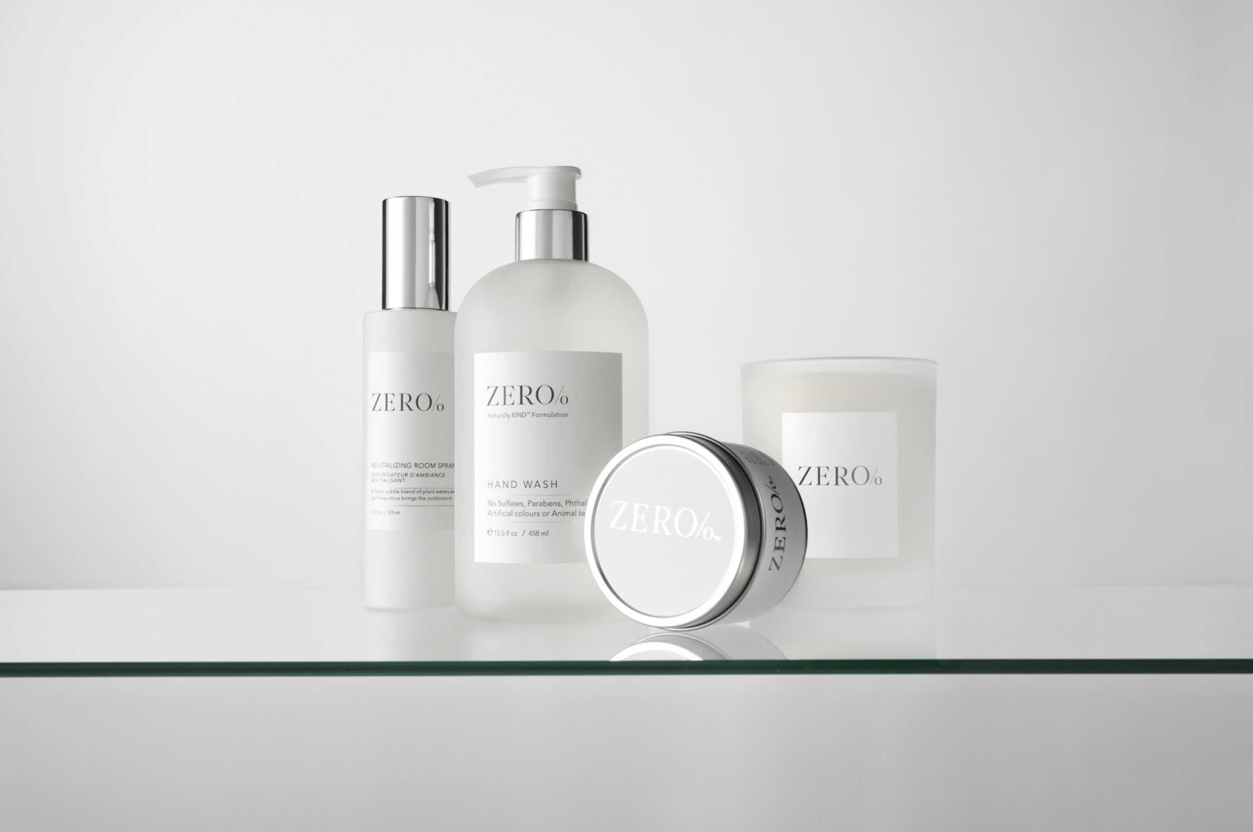 Collection of skincare and cosmetic products in white packaging with the brand name ZERO% displayed, including a spray bottle, pump bottle, jar, and candle, arranged on a clear glass surface against a white background.