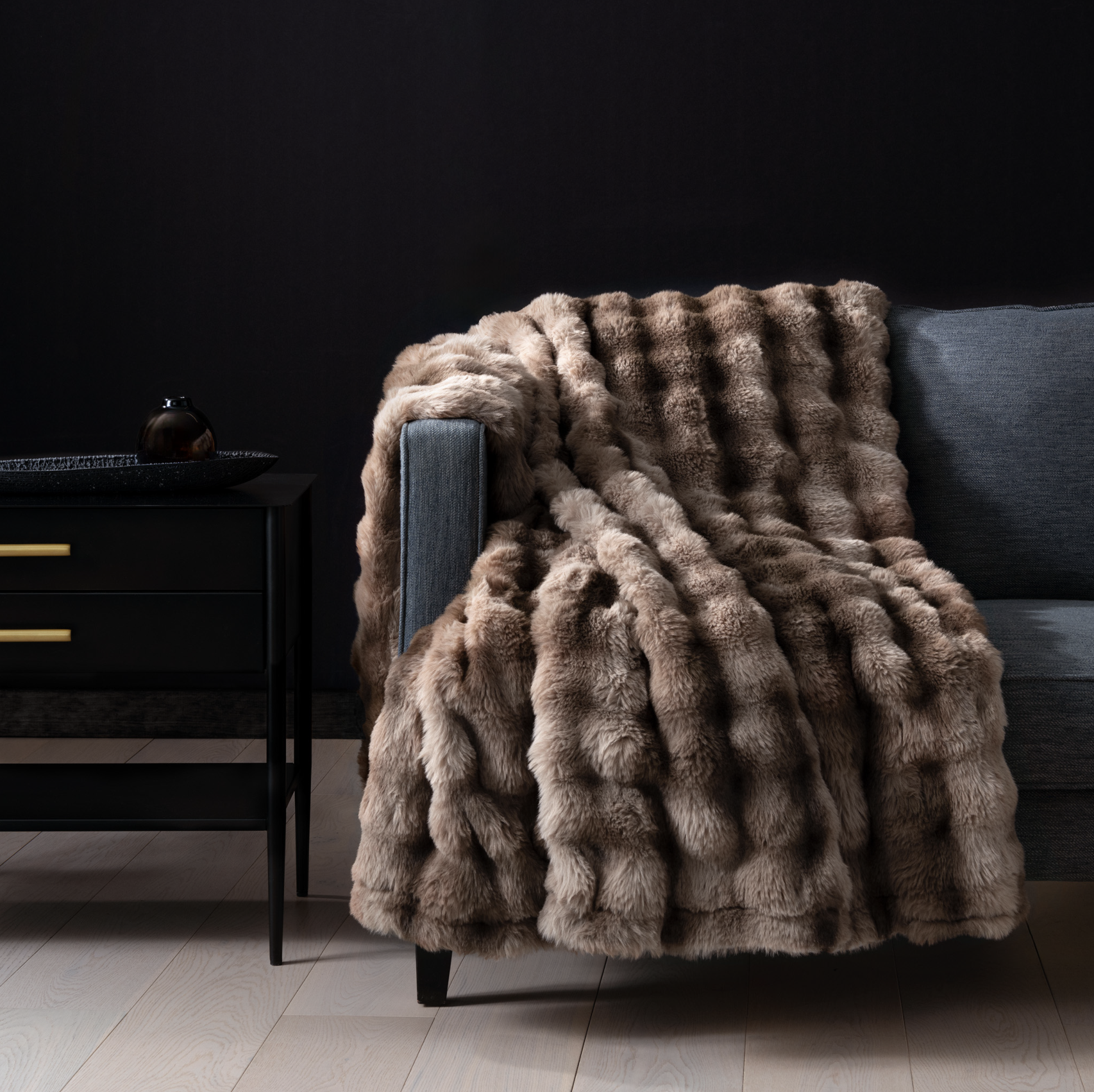A fur throw blanket draped over the arm of a dark gray sofa with a black side table and modern decor in the background.