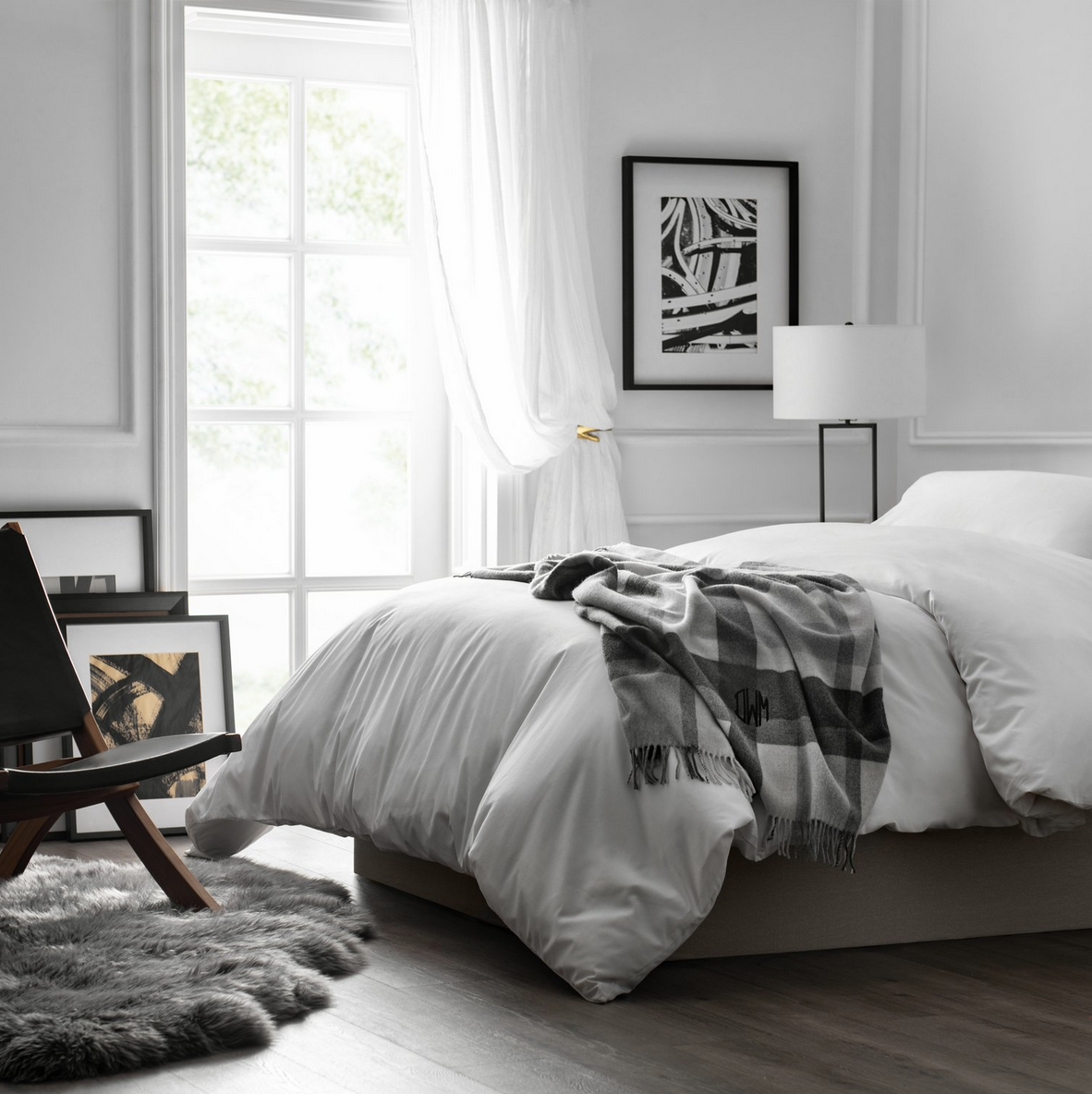 A bedroom with white walls and large window, white bed with monogrammed blanket, black and white artwork on the wall, and a wooden chair on a gray furry rug.
