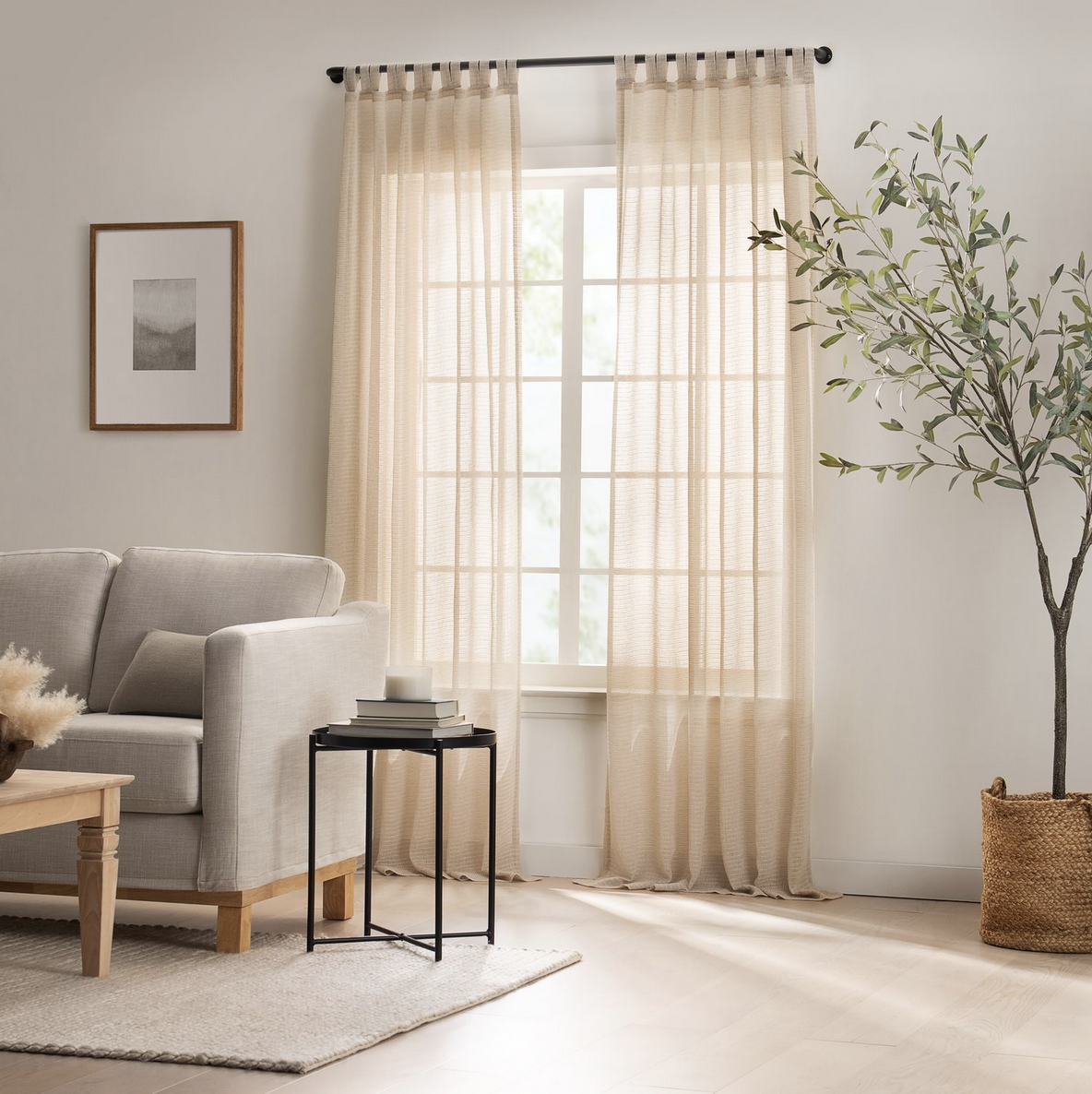 Living room with beige curtains, a light-colored sofa, a small black side table with books and a candle, a picture frame on the wall, and a potted plant in a woven basket.