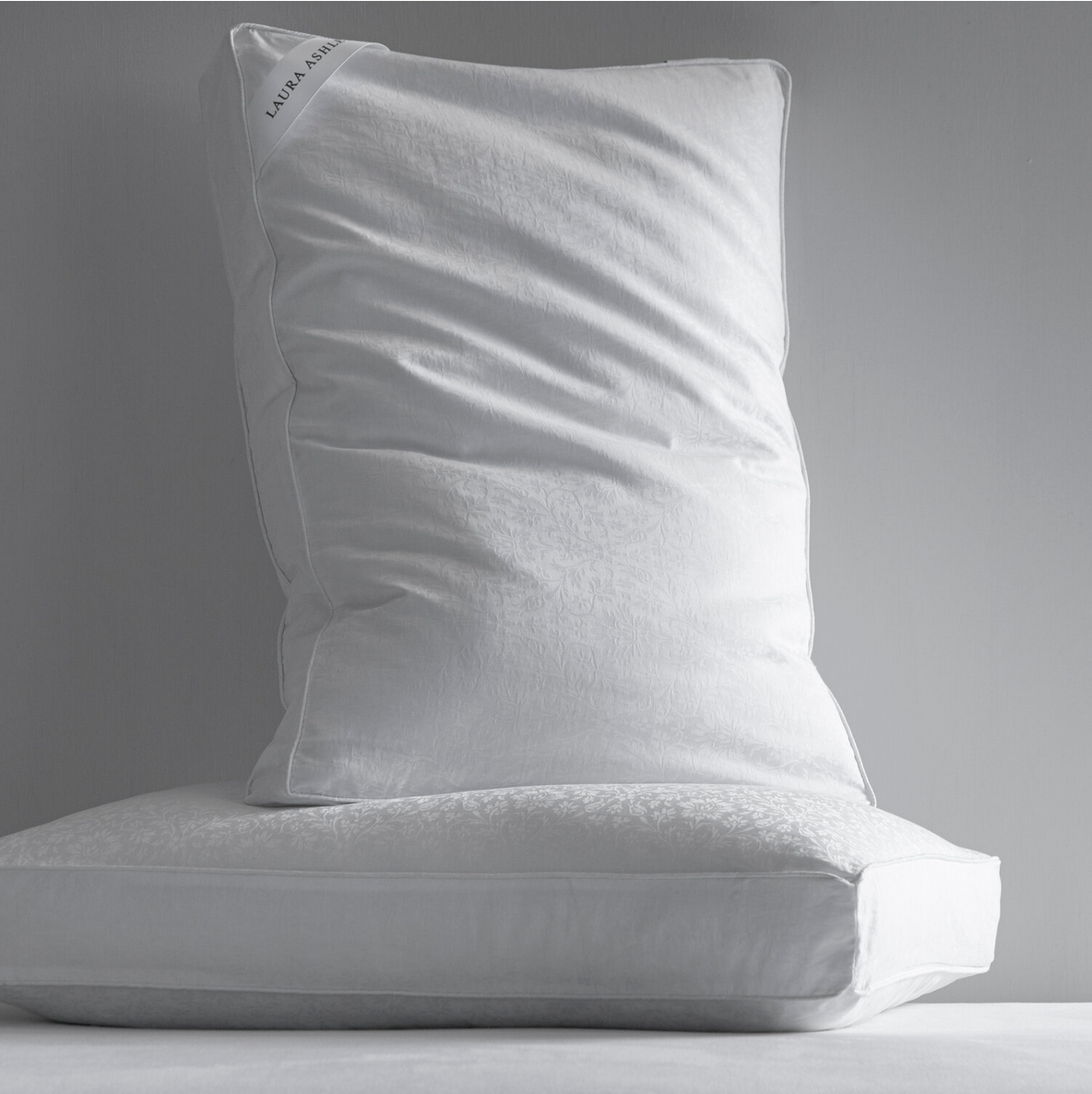 A white pillow resting on a white bed or sofa with a plain gray wall in the background.