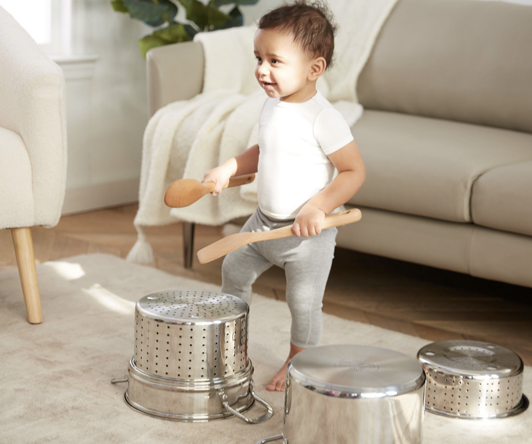 A young child playing drum on metal household items with wooden spoons in a living room.