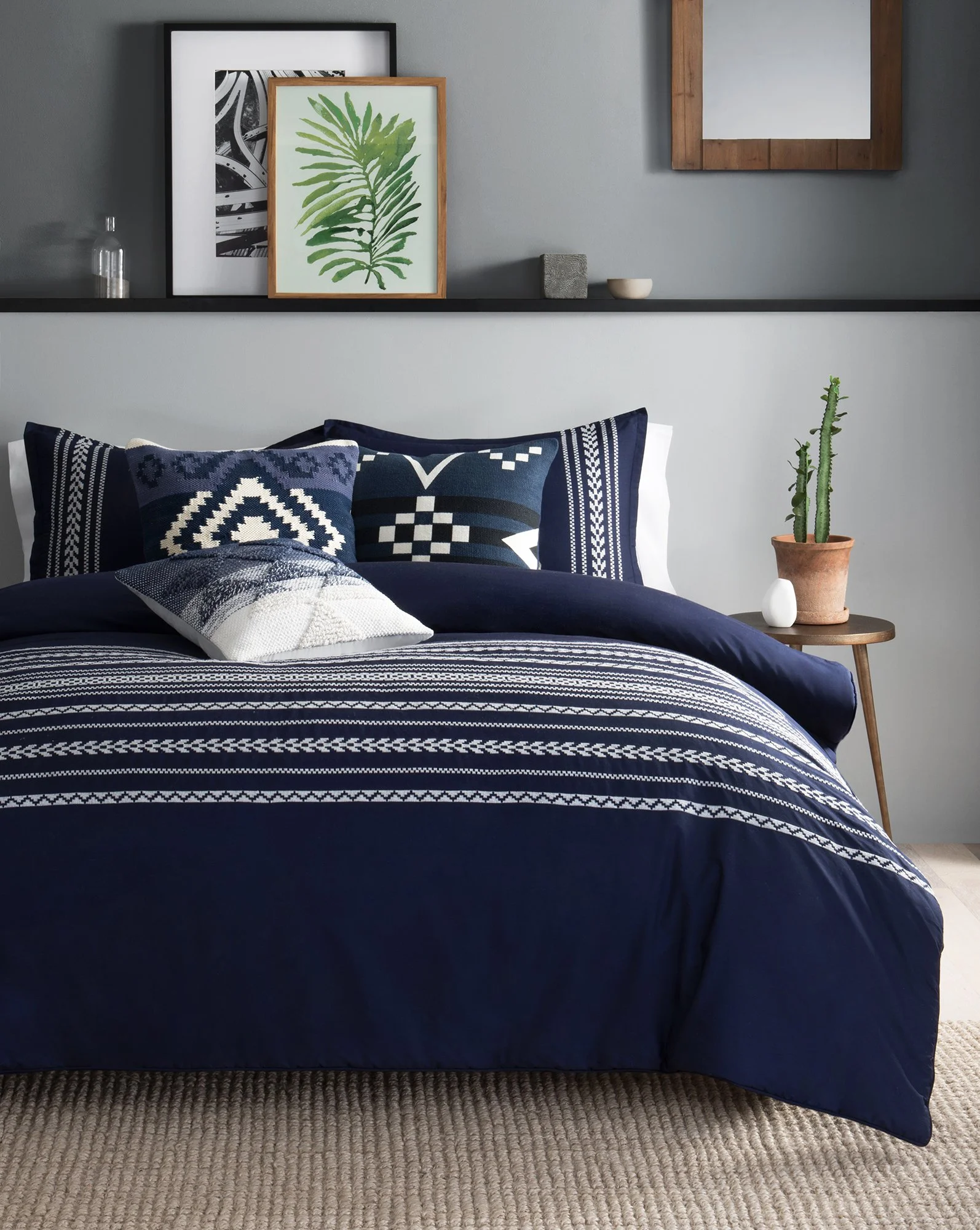A neatly made bed with navy blue and white patterned bedding, decorated with matching pillows, in a modern bedroom with gray walls, framed botanical artwork, a wooden side table with cactus plant, and wall decor.
