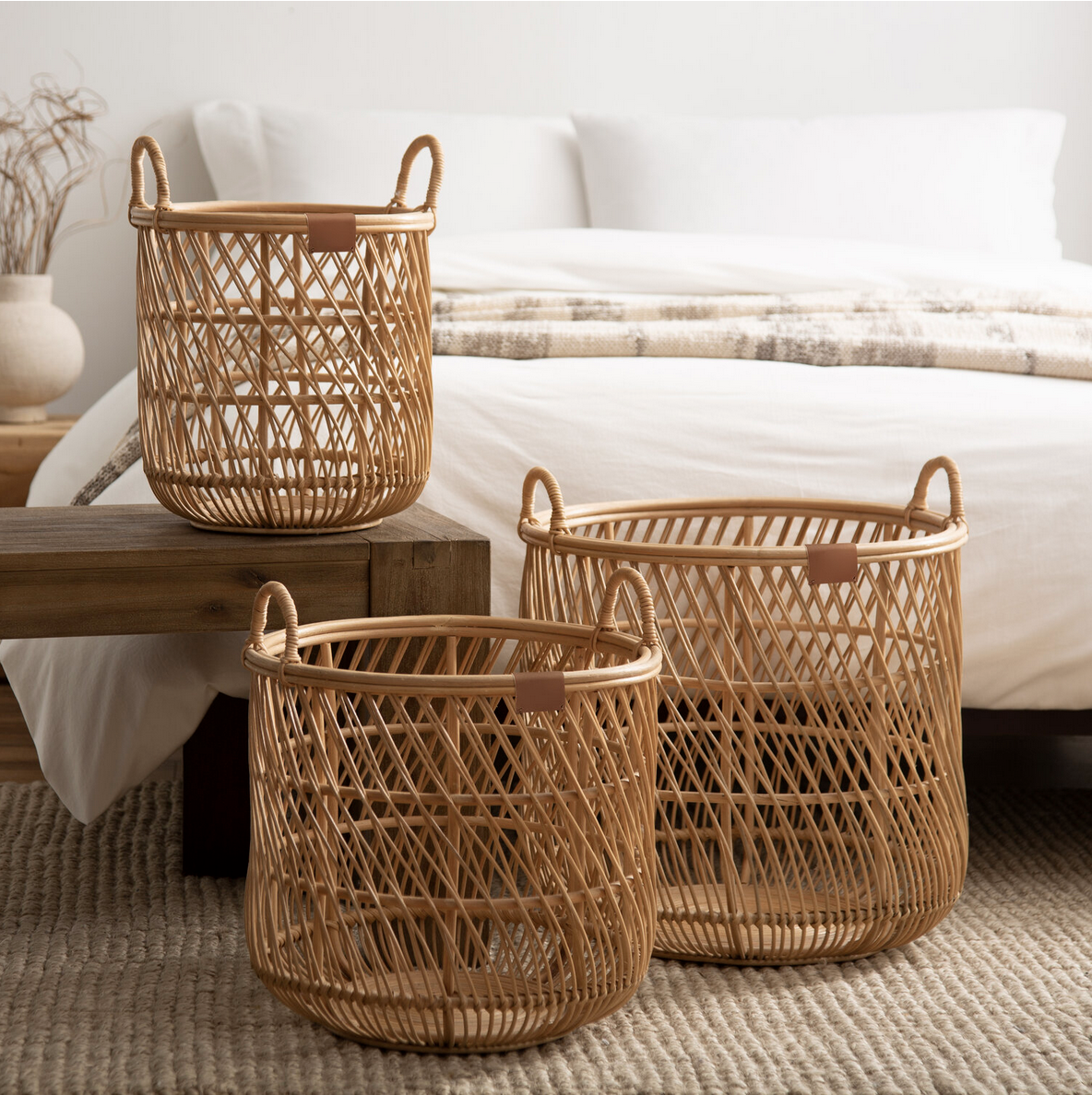 Three empty woven rattan baskets of different sizes placed on a wooden bench and on the floor near a bed in a bedroom with neutral tones.