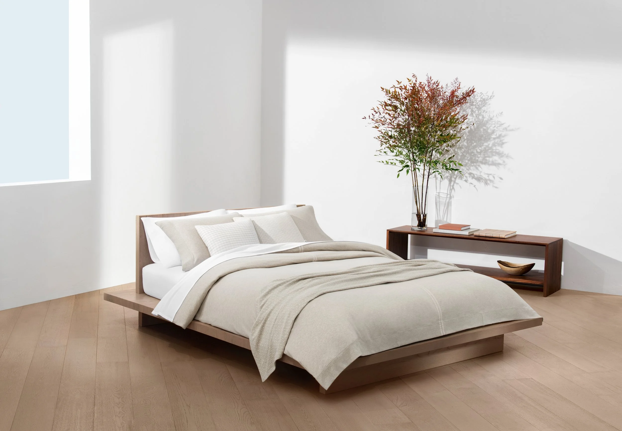 A minimalist bedroom with a low wooden bed, beige bedding, white pillows, and a wooden bench with a large vase containing branches with leaves, set against a white wall with natural light coming from a window.
