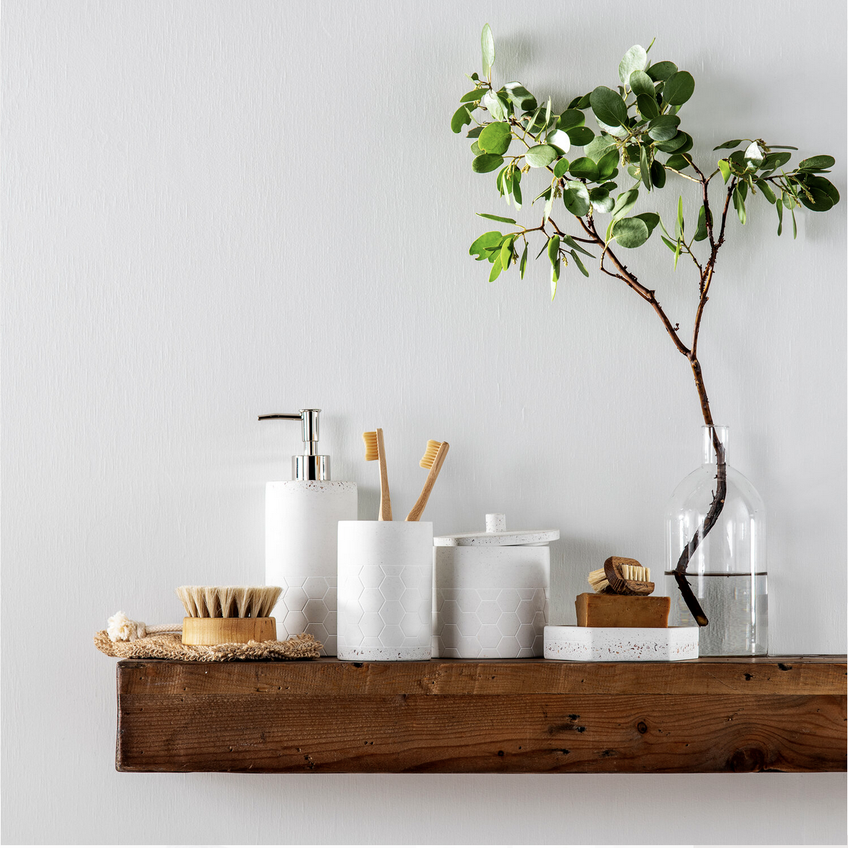 A minimalist bathroom shelf with natural wood finish, holding a white soap dispenser, two toothbrushes in a white holder, a wooden brush, a soap dish with soap, and a glass vase with a large leafy branch.