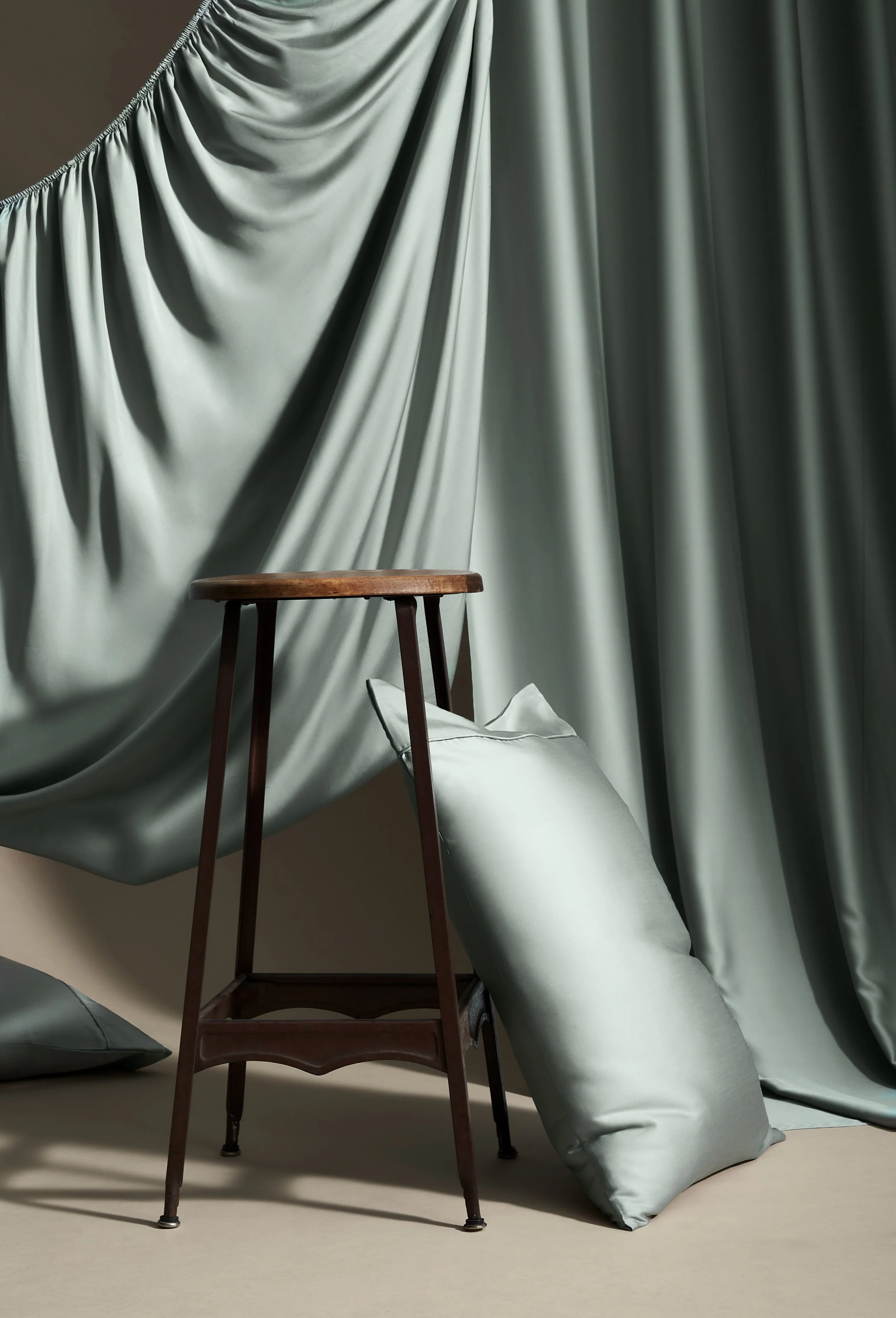 A wooden stool positioned in front of a green satin curtain and pillows, with some pillows leaning against the stool and the curtain.