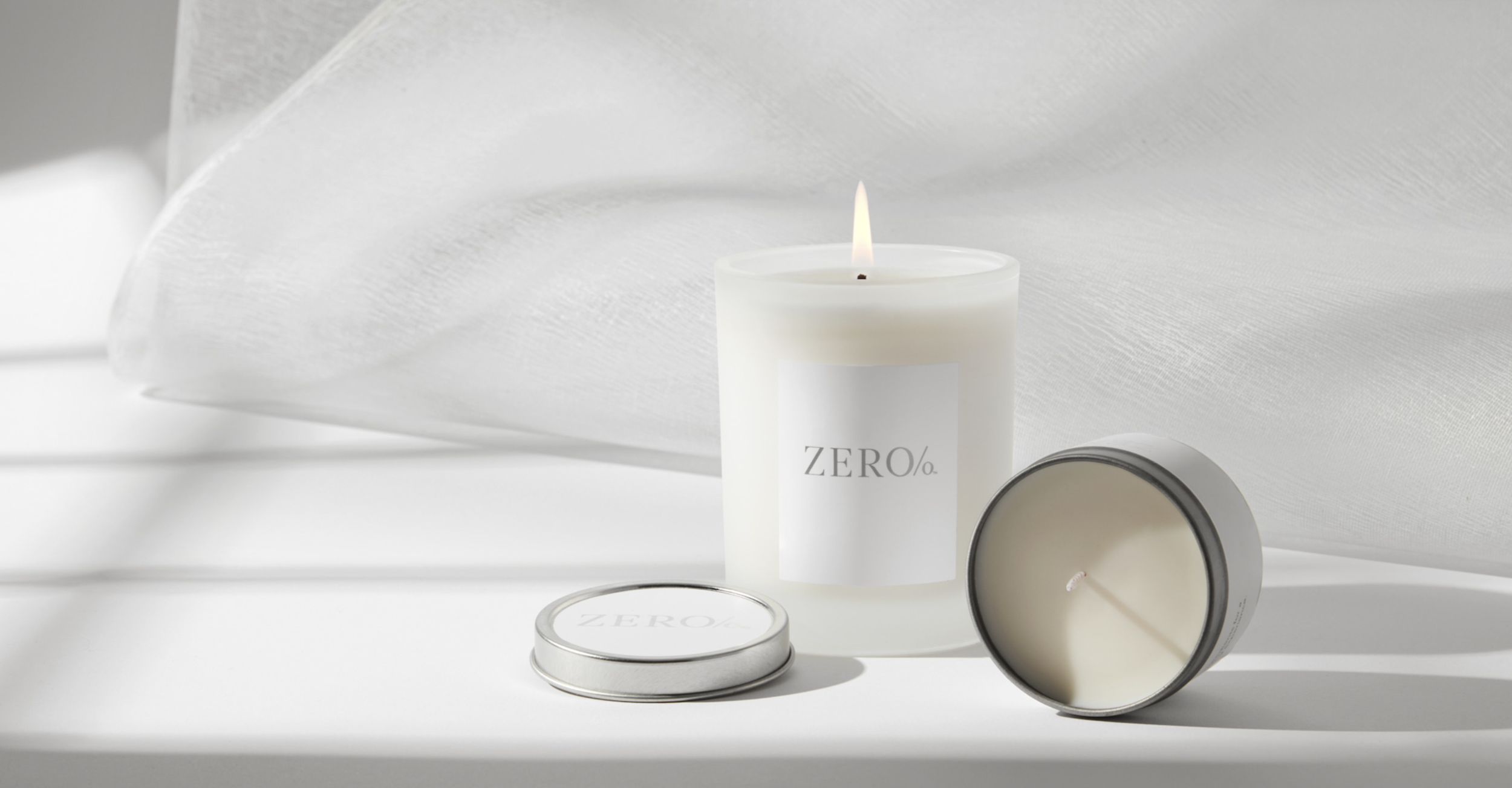 A white candle with a lit flame and a label that reads 'ZERO/' is placed on a white surface. Two metallic candle tins are also present, one upright and the other lying on its side showing the unused wick, all against a textured white background.