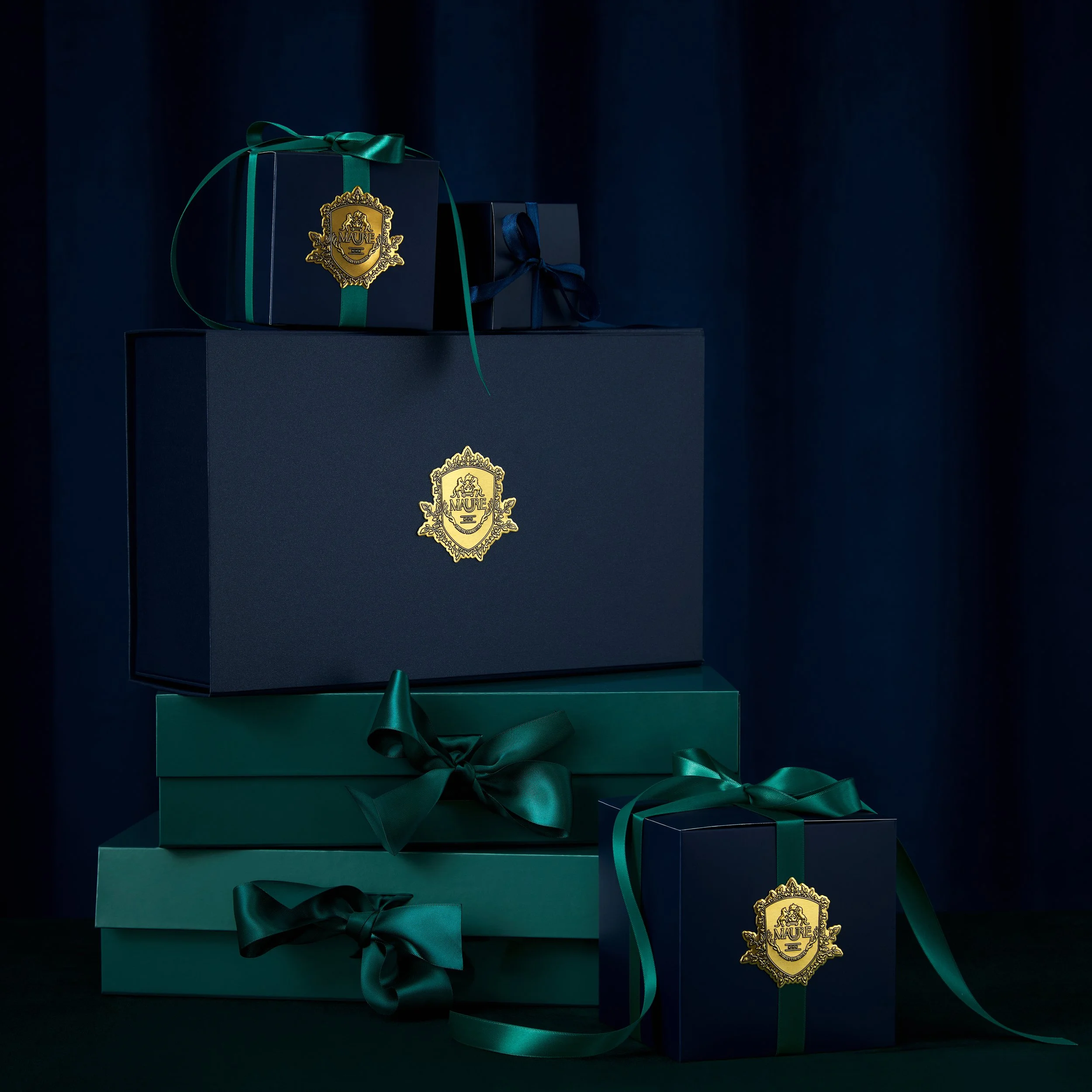 Stack of elegant gift boxes in dark blue and green with satin ribbons and gold crest logos against a dark background.