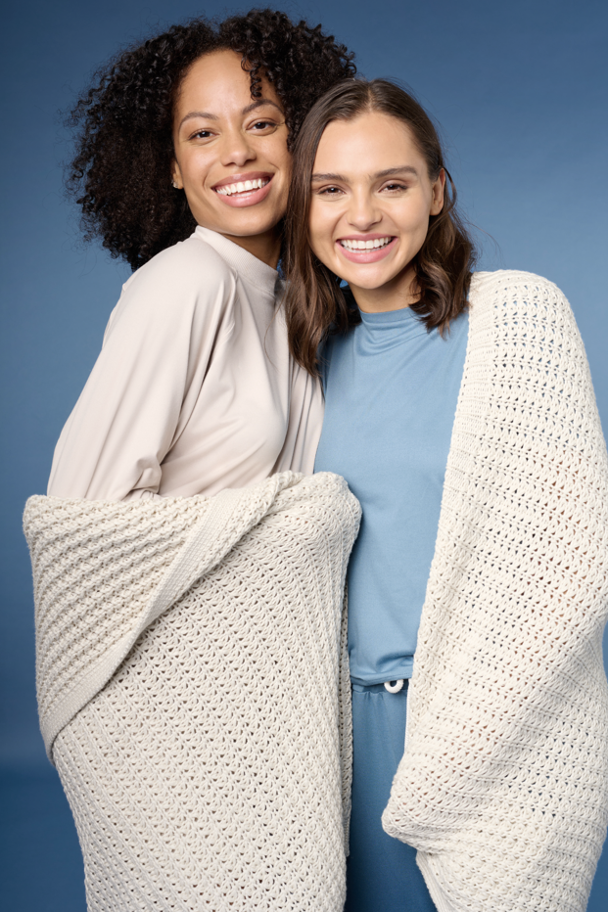 Two women smiling and standing close, wearing cozy sweaters, with a blue background.