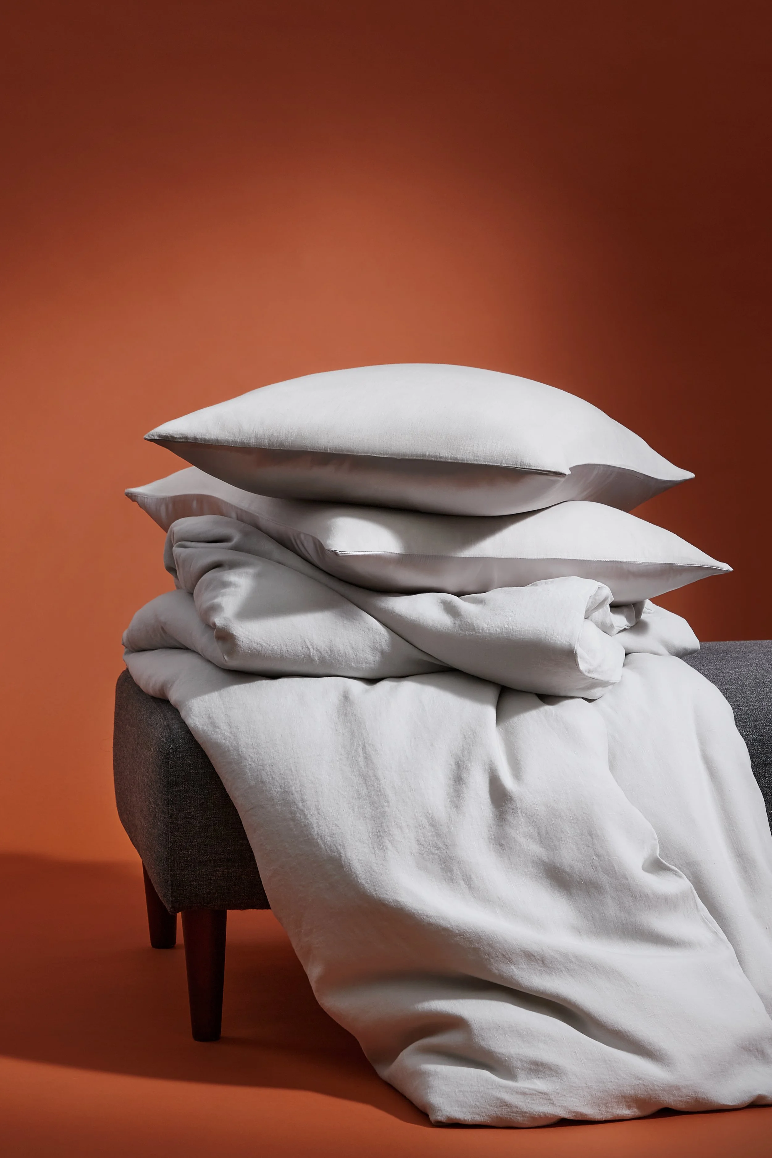 A pile of white pillows and blankets on a grey upholstered bench against an orange wall.