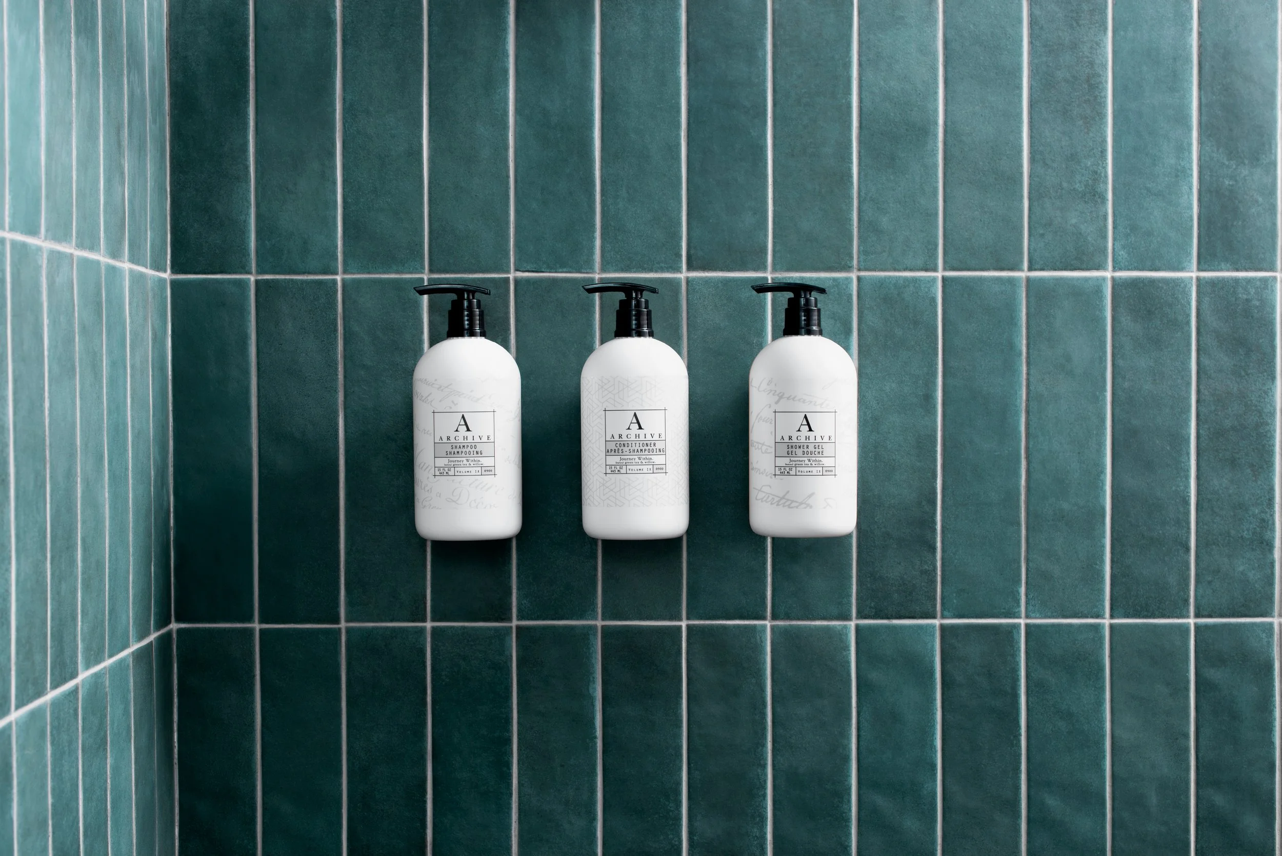 Three white bottles of shampoo, conditioner, and shower gel mounted on a teal tiled bathroom wall with black pump dispensers.
