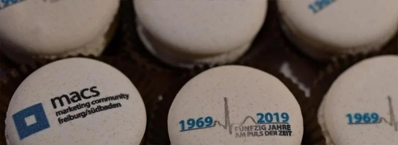 Weiße Kugeln mit Texten und Logos, einige heißen 'macs' mit dem Schriftzug 'marketing community freiburg/sulzbach' und andere zeigen die Jahreszahlen 1969-2019 mit einer Herzmonitor-Linie und dem Text 'FÜNFZIG JAHRE AM PULS DER ZEIT'.