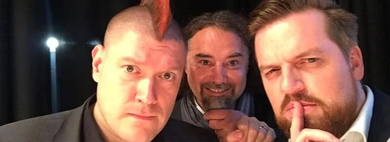 Drei Männer, die für ein Selfie posieren, einer macht ein Geheimniszeichen mit dem Finger vor den Lippen, alle schauen in die Kamera.