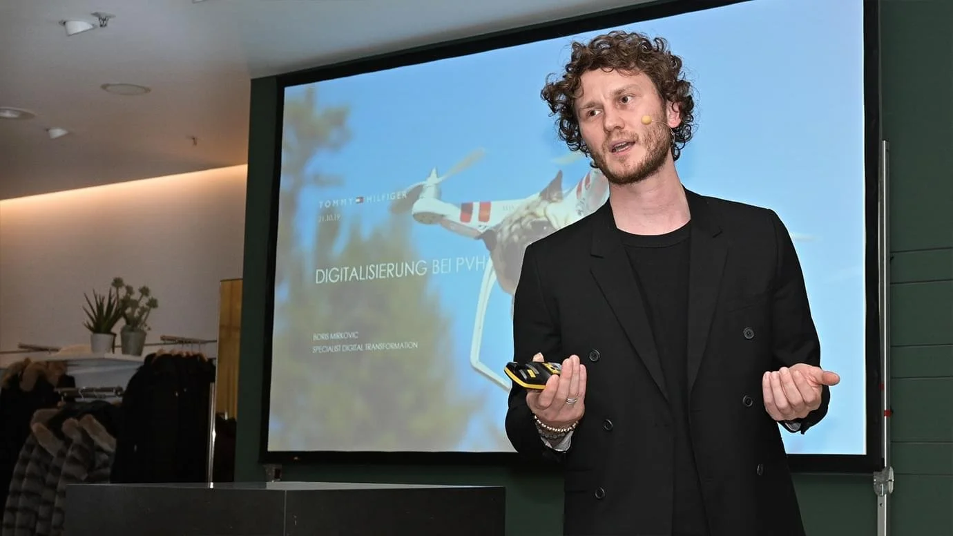 Mann hält Präsentation vor einem Bildschirm mit Flugzeug und Text auf Deutsch.