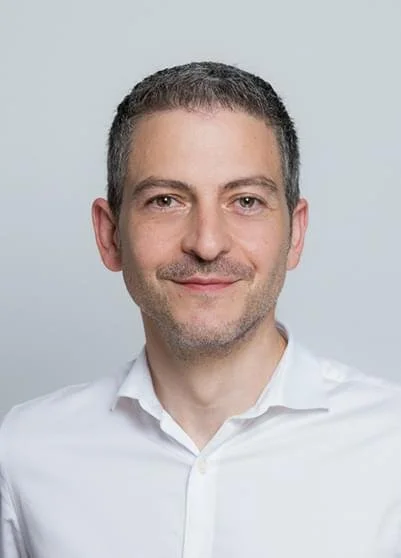 Porträt eines Mannes mit kurzen braunen Haaren, leichtem Bart, in einem weißen Hemd, vor heller Hintergrund.