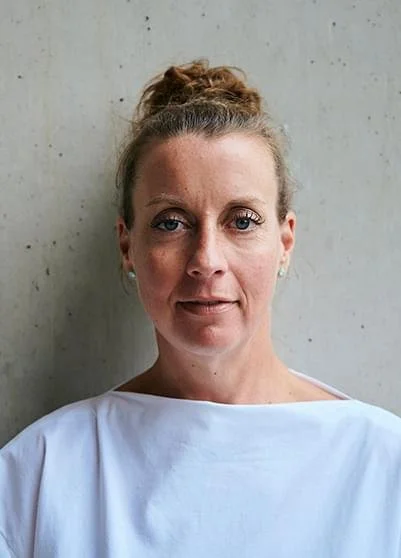 Porträt einer Frau mit hellbraunen Haaren in einem lockeren sogenannten Topknot, schwarzer Oberbekleidung und stehend vor einer grauen Betonwand.